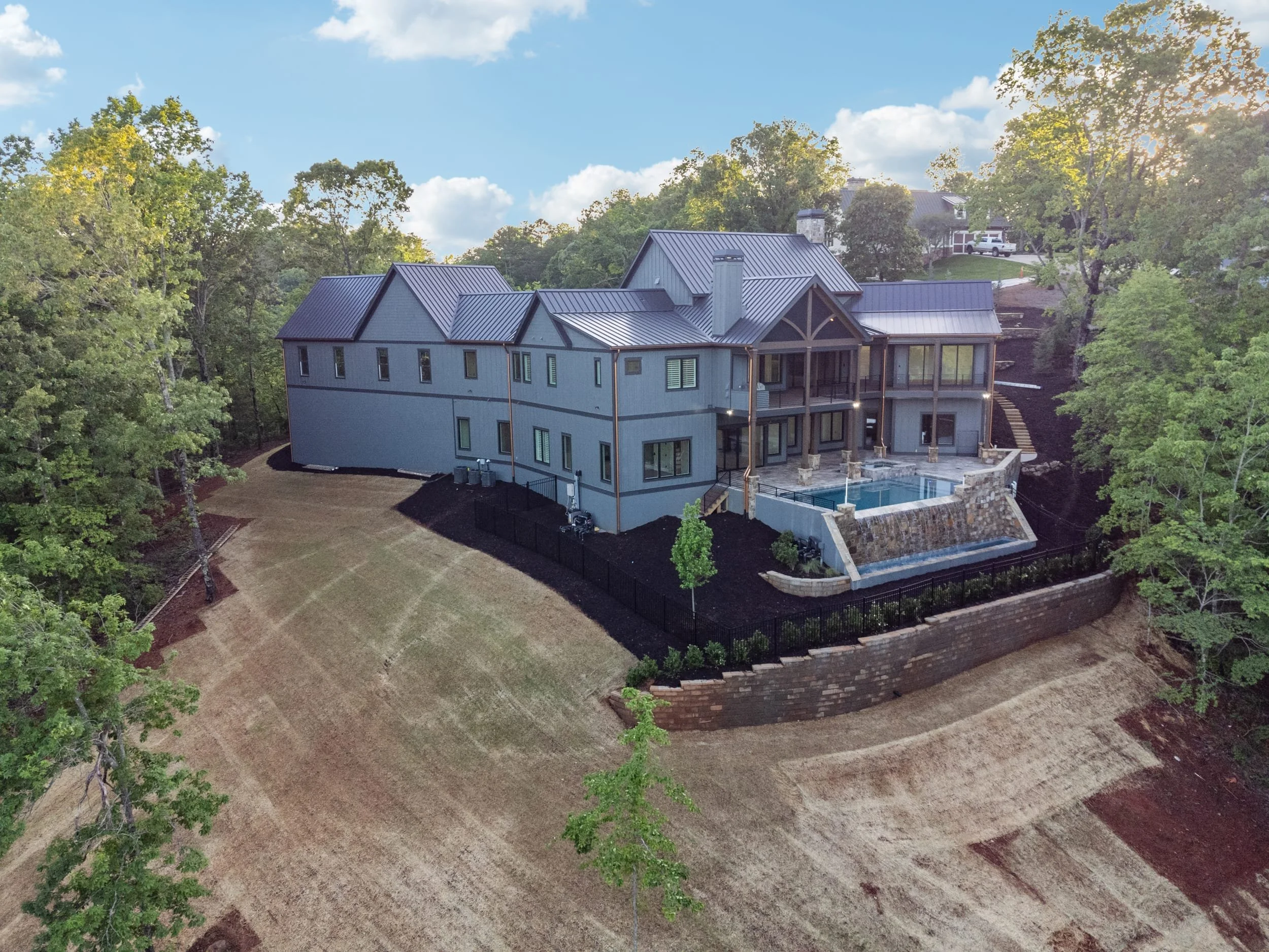 A large modern house with multiple roof peaks, blue exterior walls, and a backyard swimming pool surrounded by a stone wall, located on a hillside amid lush green trees under a partly cloudy sky.