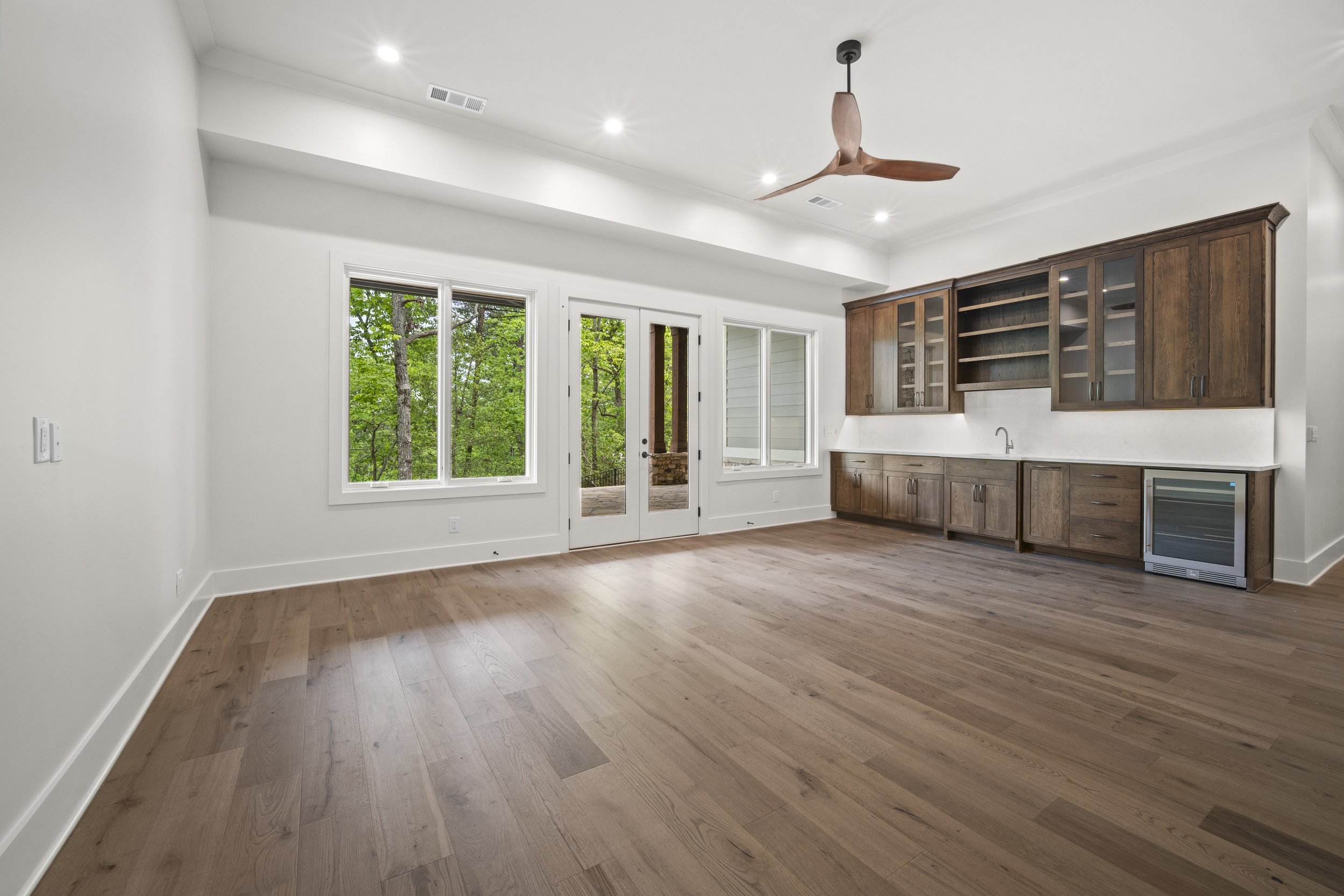 Empty living space with hardwood flooring, white walls, large windows showing greenery outside, and built-in wooden cabinets.