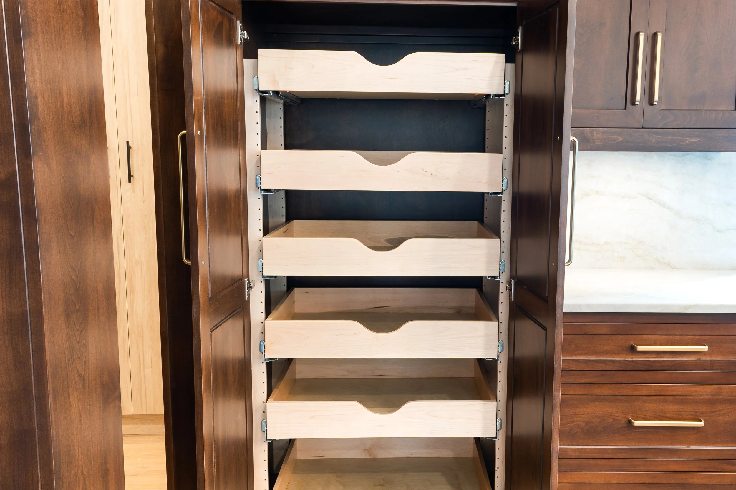 Open kitchen cabinet with three pull-out shelves made of light wood, surrounded by darker wood cabinets with gold handles.