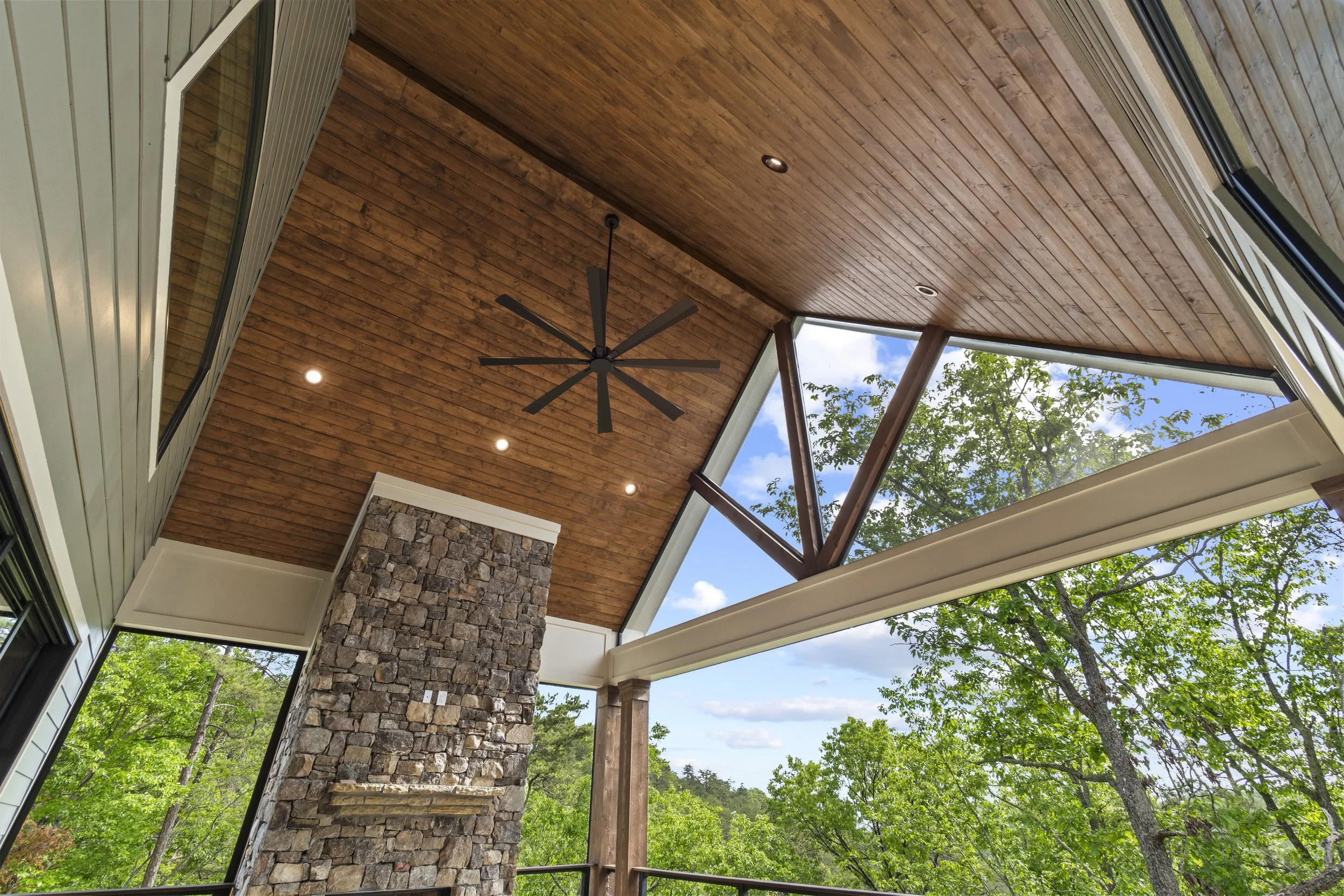 A view of a covered patio with a wooden ceiling, a ceiling fan, recessed lighting, large windows, and a stone chimney, with trees and a blue sky with clouds outside.