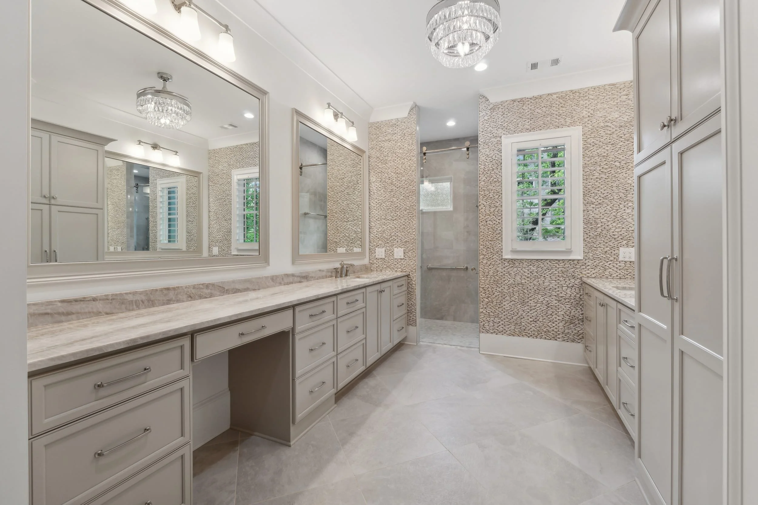 A spacious bathroom with white cabinets, a marble countertop, large mirrors, a shower with glass doors, and two large windows with white shutters.