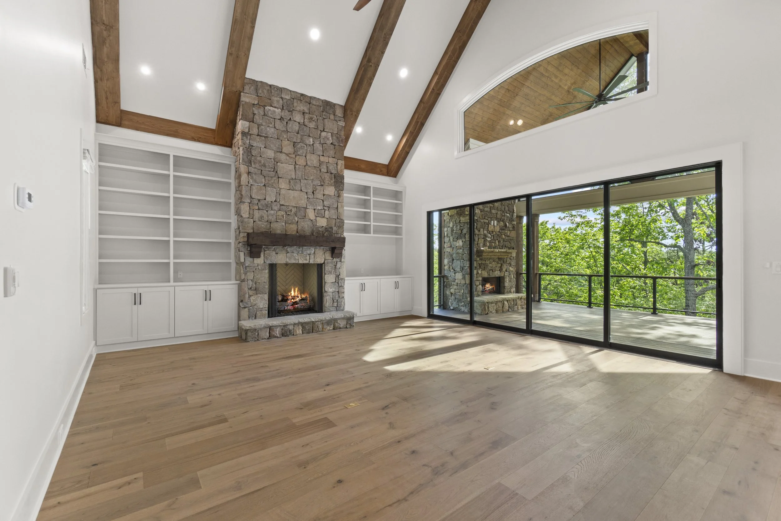 Interior of a modern living room with a stone fireplace, built-in white shelves, wood beam ceiling, large sliding glass doors leading to an outdoor deck with a stone column and trees visible outside.