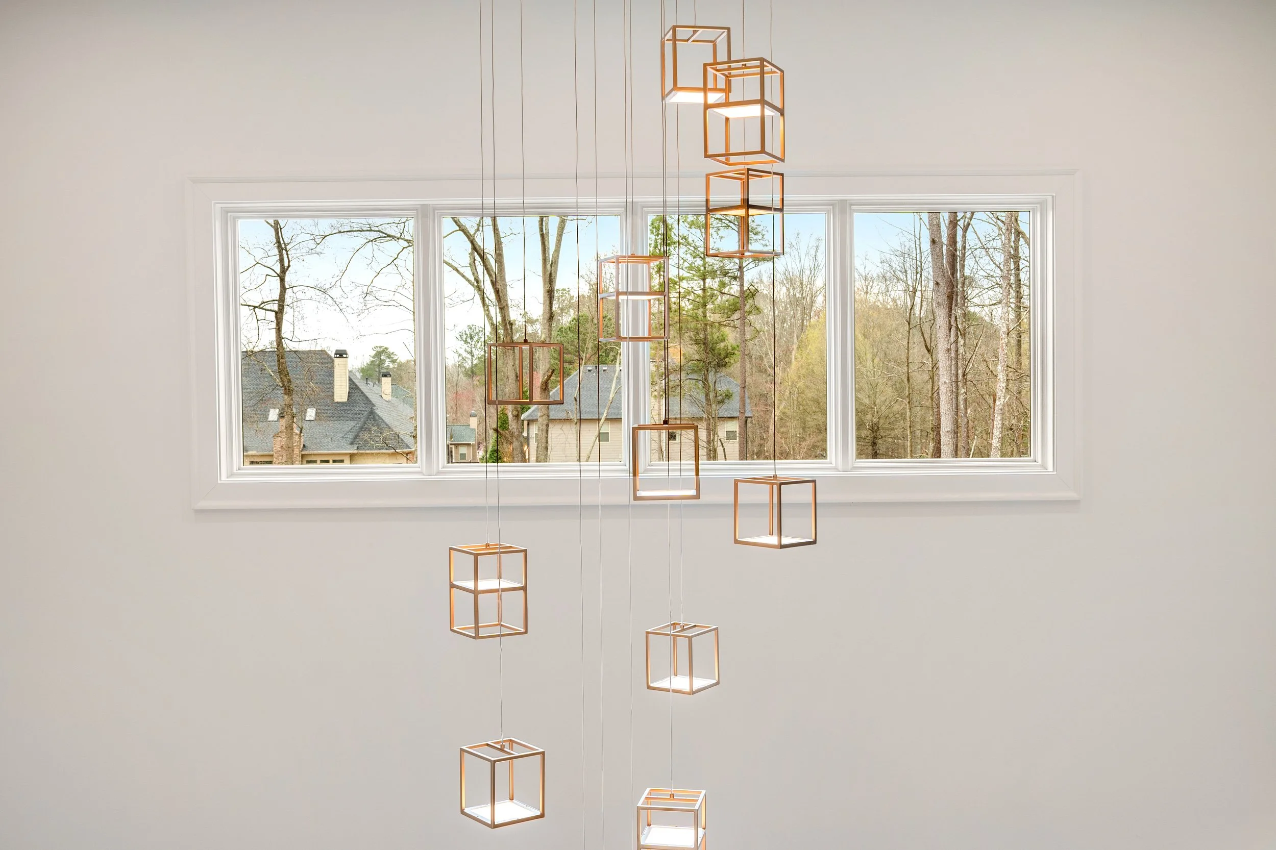 Modern geometric chandelier hanging in front of a large window with trees and rooftops visible outside.