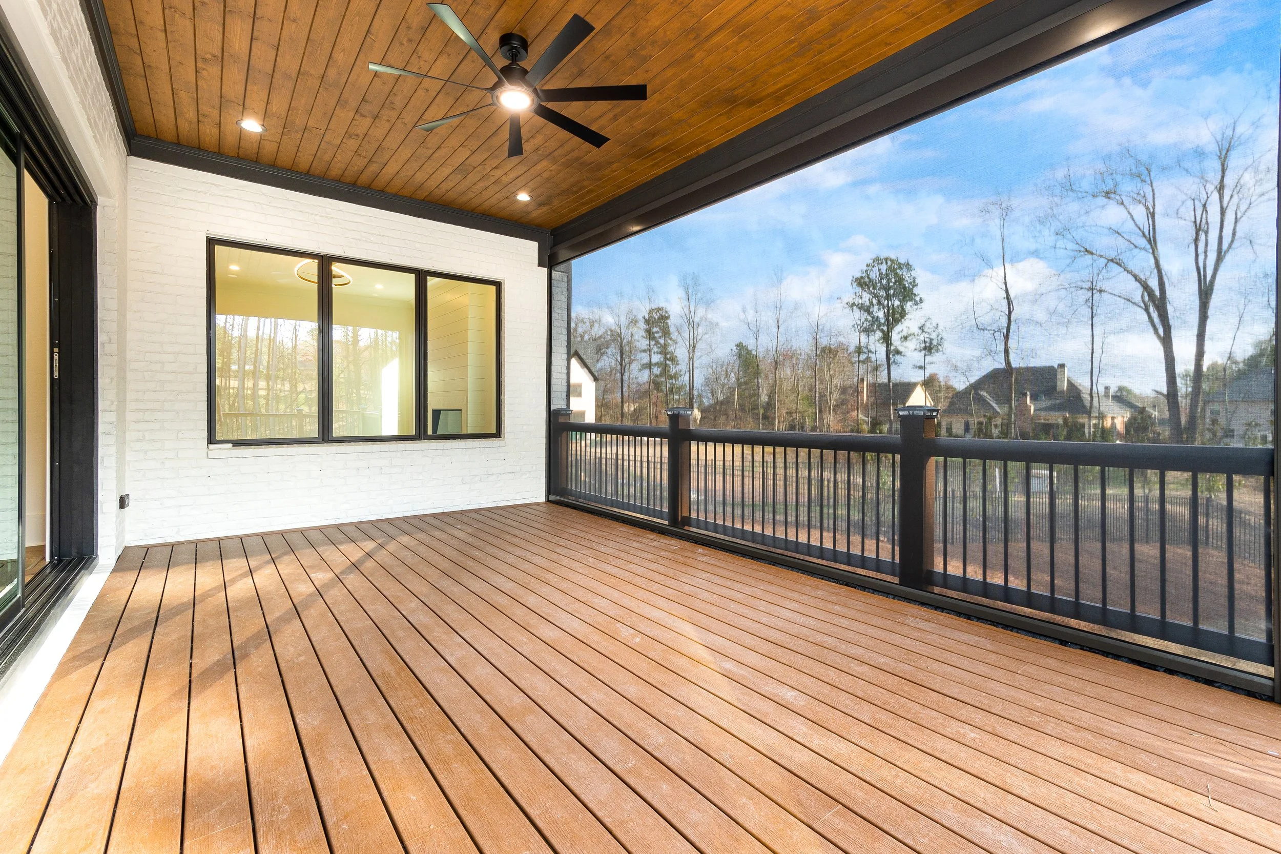 An outdoor balcony with a wooden floor, black railing, white brick wall, window, ceiling fan, and a view of trees and neighboring houses under a blue sky.