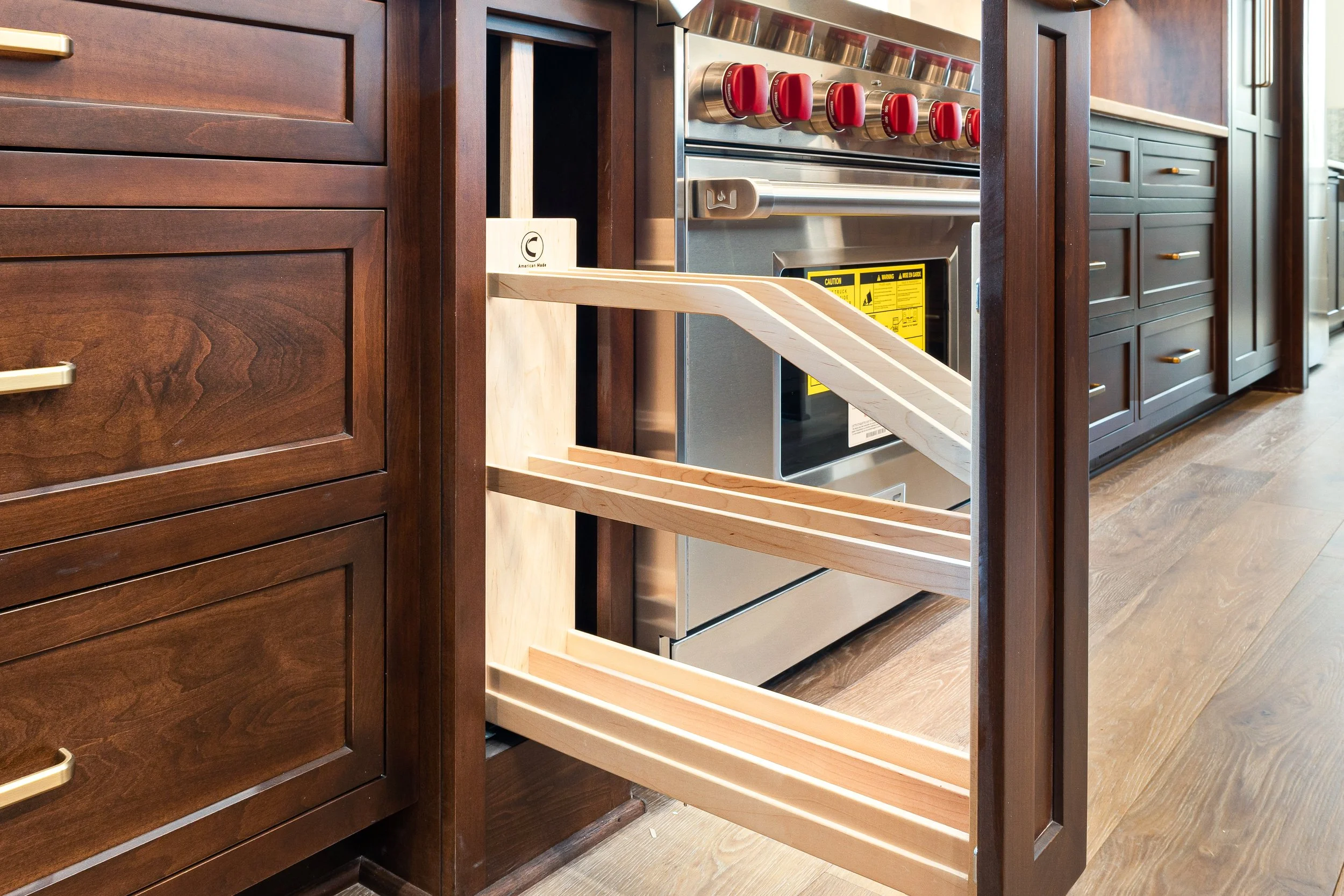Unfinished kitchen cabinet with a pull-out drawer, located between dark wood cabinets and wooden flooring.