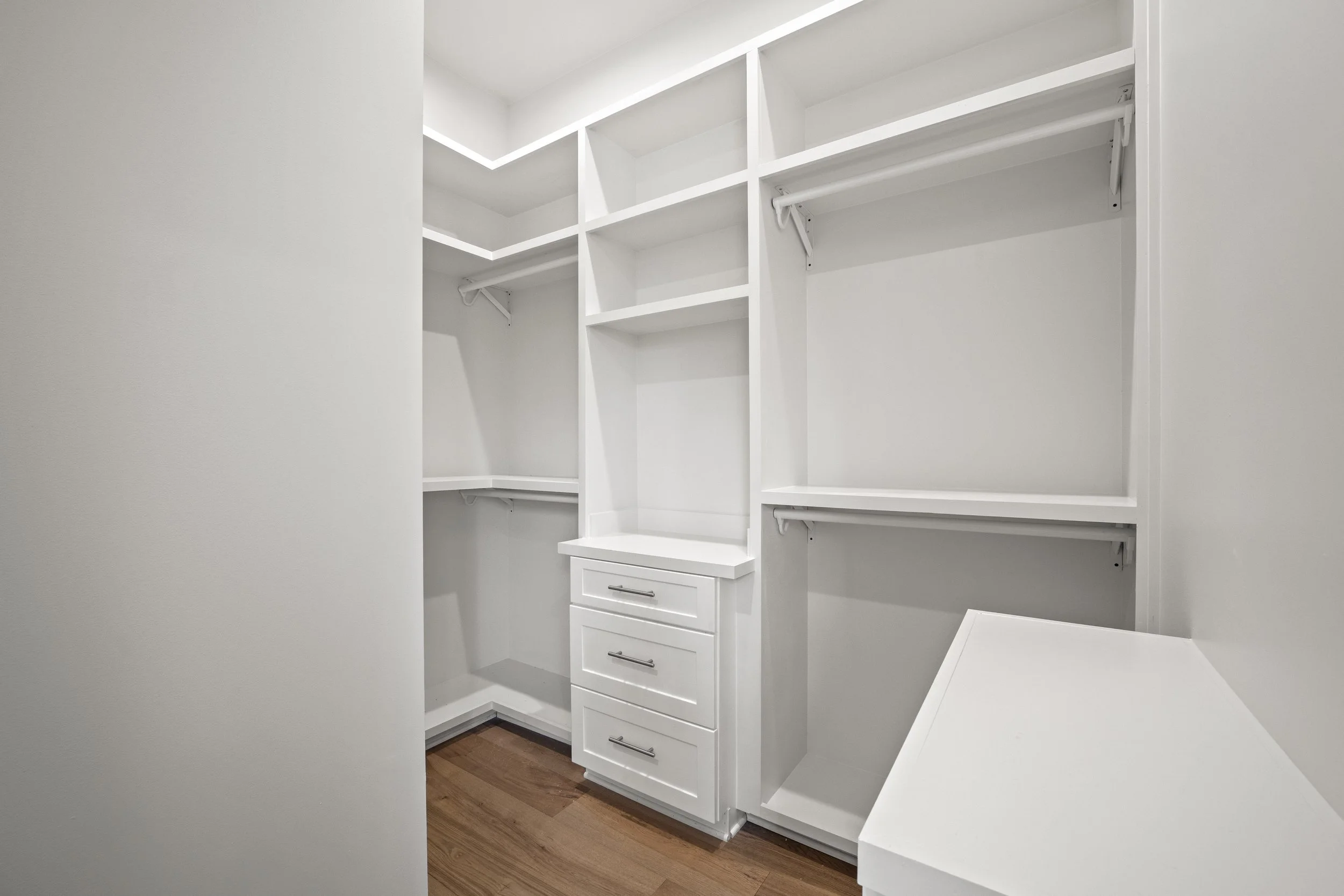 Empty white walk-in closet with shelves, drawers, and hanging rods, with wooden flooring.