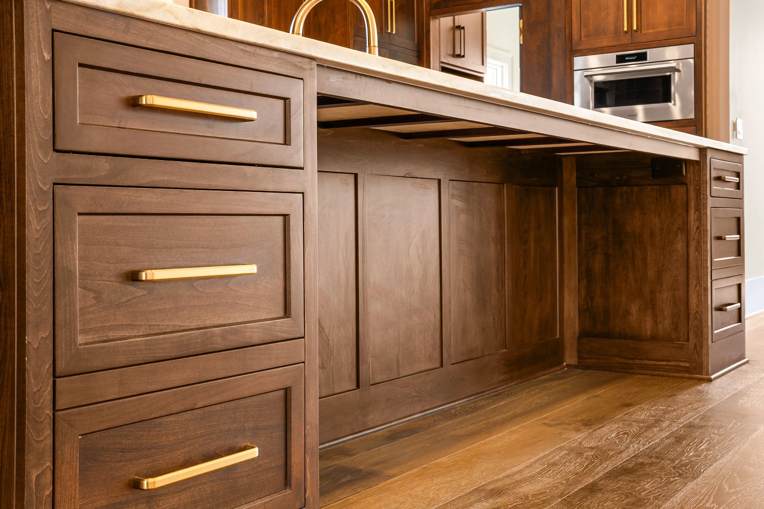 Close-up view of a kitchen countertop, cabinets, and drawers made of dark wood with gold hardware, in a kitchen with built-in appliances and hardwood flooring.