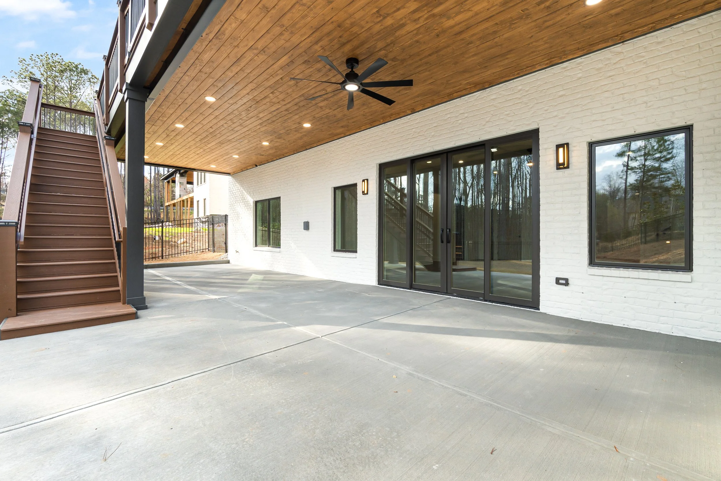 The underside of a covered patio or balcony with wooden ceiling, ceiling fan, recessed lights, black outdoor wall lights, and sliding glass doors leading inside. There are stairs on the left side of the image, and a concrete ground surface with parki