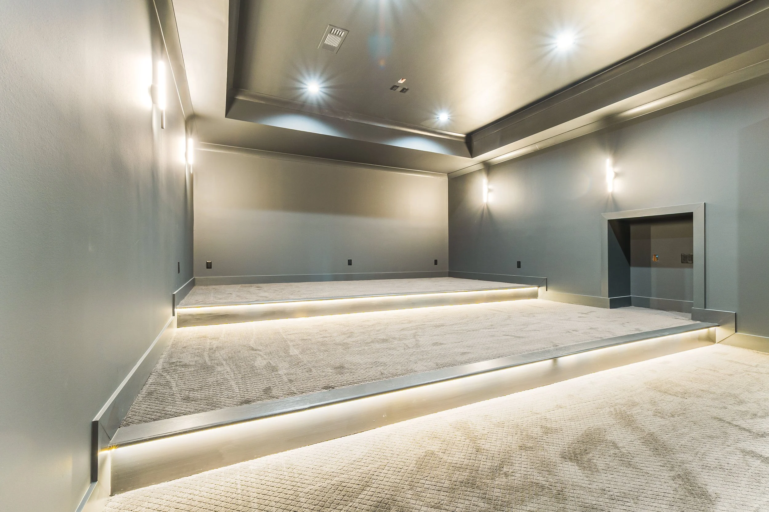Empty, modern interior space with carpeted floors, gray walls, and a tray ceiling with inset lighting. There are steps leading up to a slightly elevated platform with concealed lighting underneath.