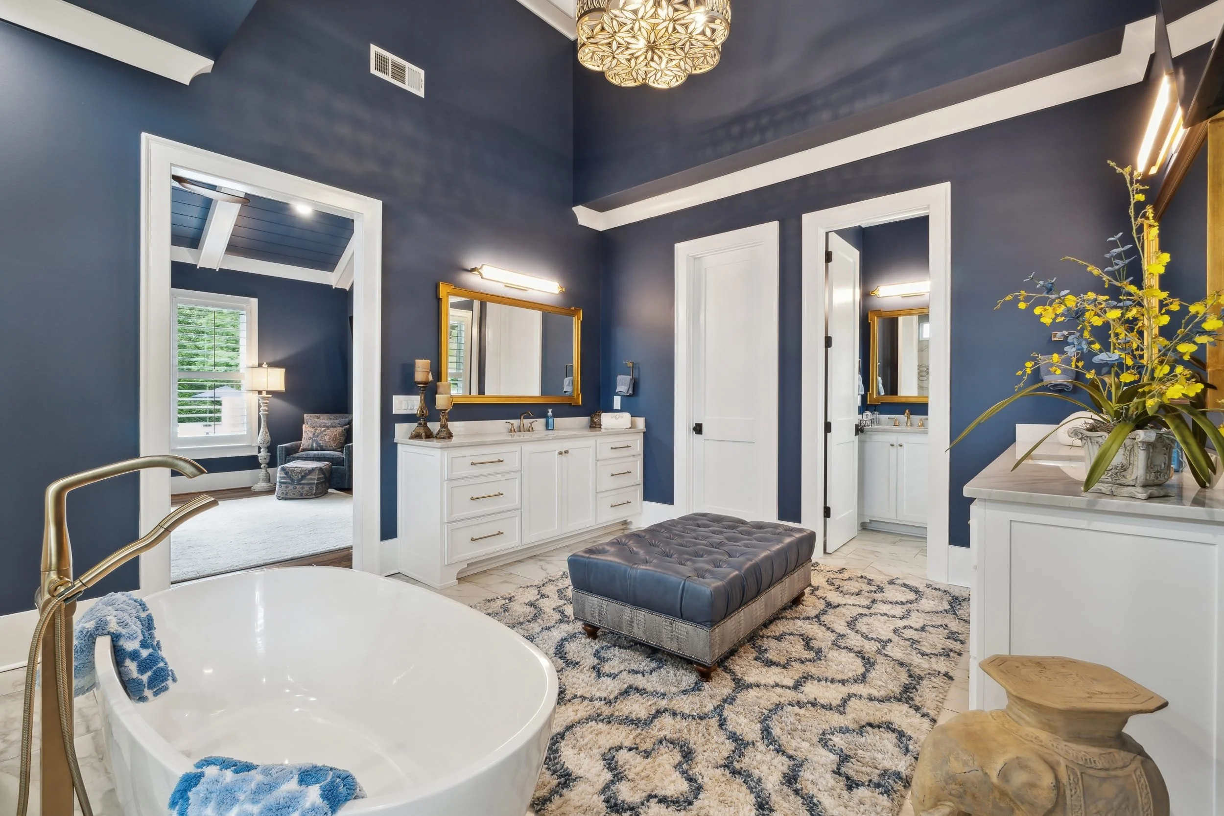 A bathroom with navy blue walls, white trim, and a large mirror with a gold frame. There is a white vanity with gold handles, a tufted bench, a decorative rug, and a flower arrangement on a white counter. A partial view of a bathtub with a gold faucet and a blue towel can be seen in the foreground. There's also a sitting area with a lamp and window in the adjacent room.