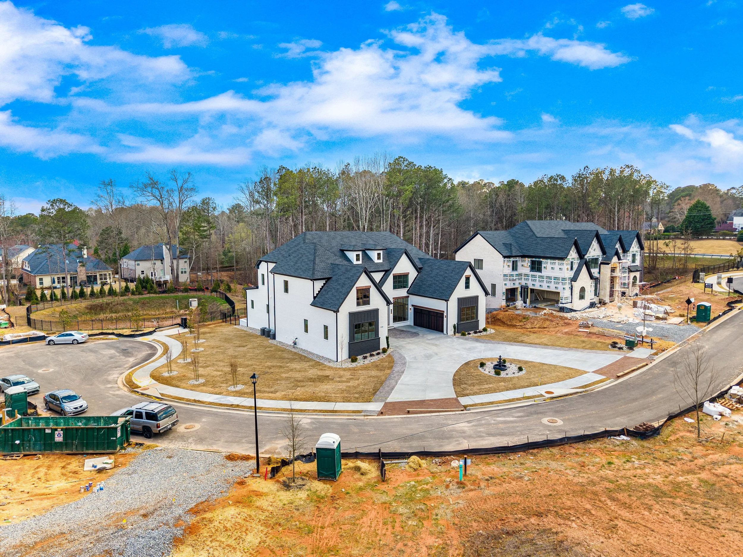A suburban neighborhood under construction with several houses, some of which are still being built. There are parked cars, a circular driveway, and a grassy area with young trees. The sky is partly cloudy with patches of blue.