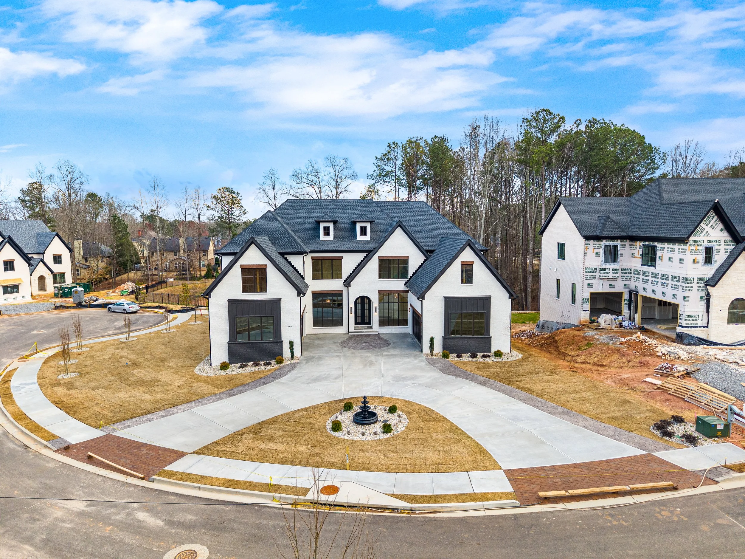 A newly constructed white house with black roof accents in a suburban neighborhood. The front yard has a circular driveway, grass, and a small fountain at the center. There is ongoing construction on neighboring houses, and trees in the background un