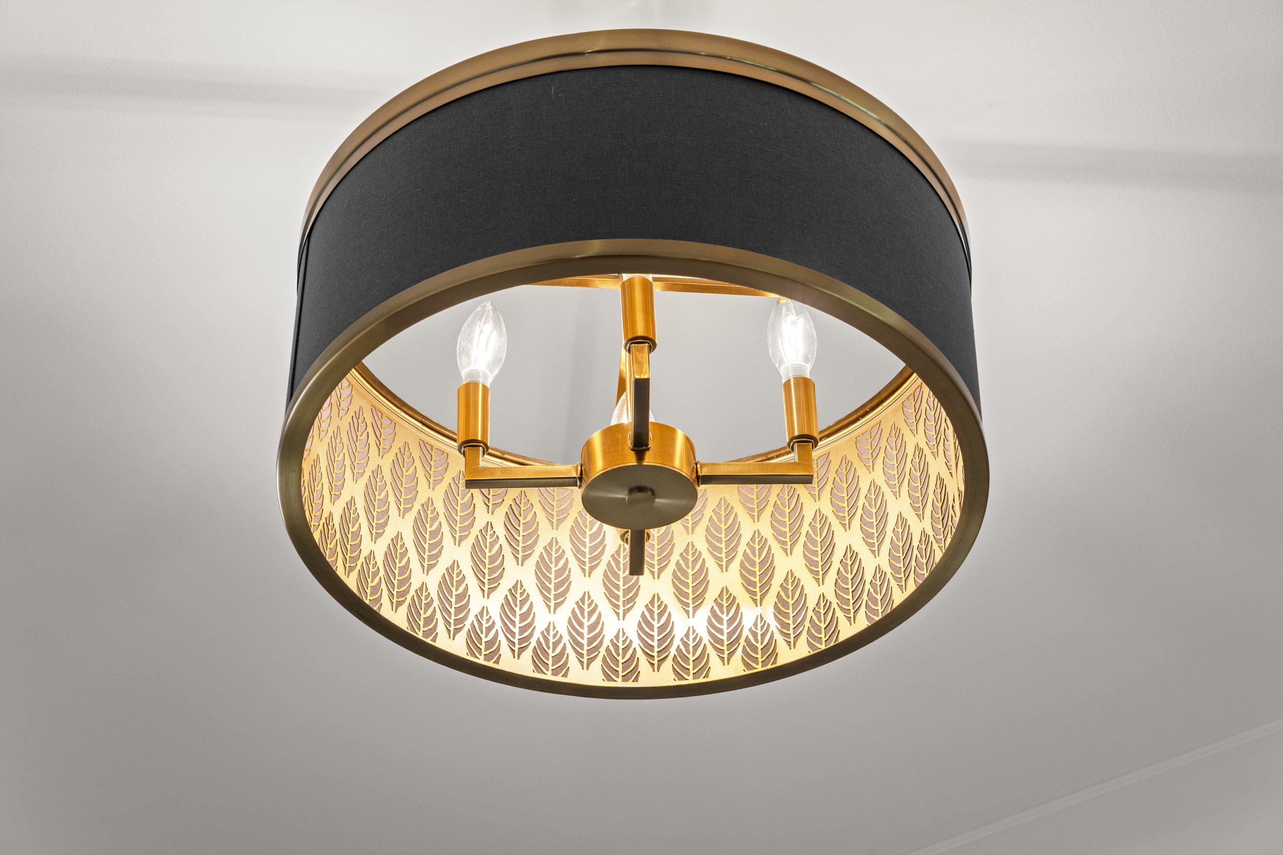 Ceiling light fixture with a black, rounded shade and a gold decorative band, featuring a patterned inner shade with a leaf motif, mounted on a white ceiling.