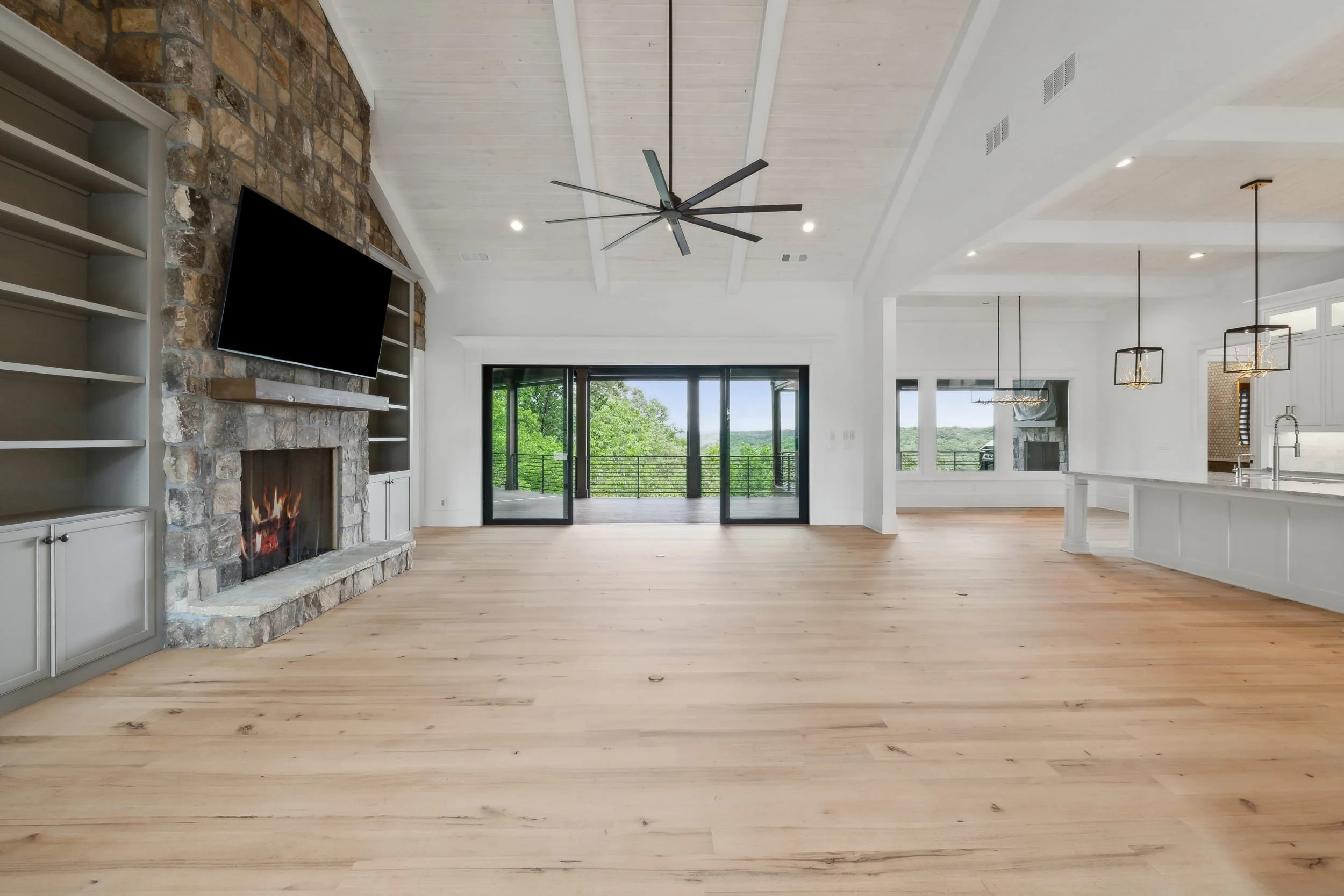 Open-concept living room and kitchen with light wooden flooring, white walls, a stone fireplace, a wall-mounted TV, large sliding glass doors leading to a balcony, and modern light fixtures.