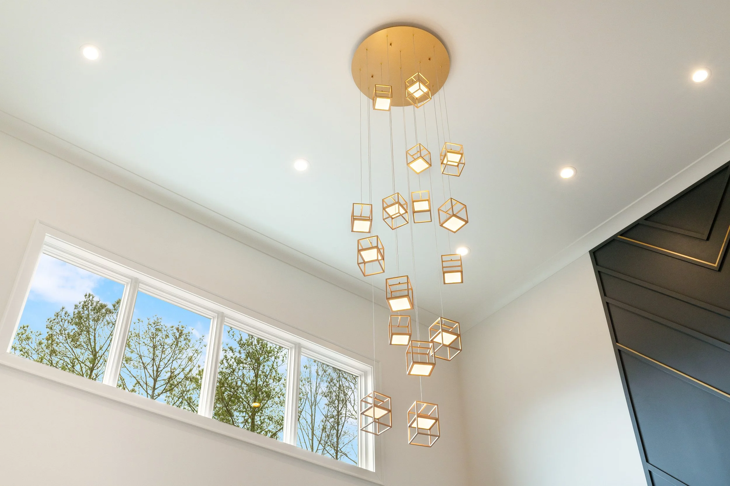 Modern chandelier with geometric cube-shaped lights hanging from a round ceiling mount in a bright room with large windows showing trees outside.