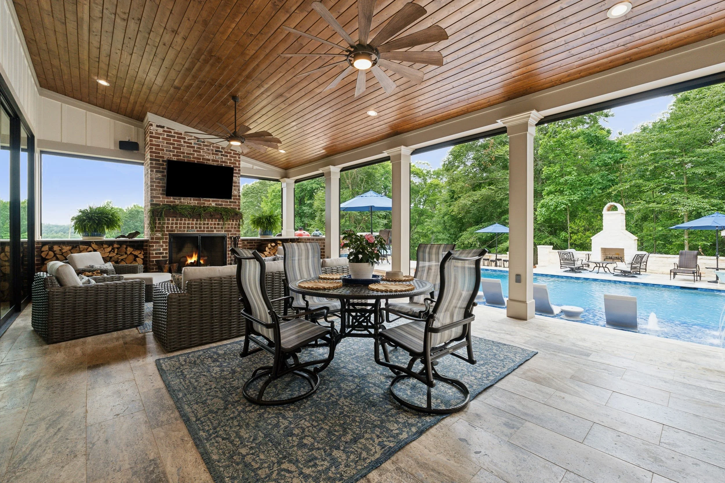Covered outdoor patio area with wicker seating, a round dining table with striped chairs, a brick fireplace with a television mounted above, ceiling fans with wooden blades, and a view of a backyard swimming pool with patio furniture and umbrellas surrounded by trees.