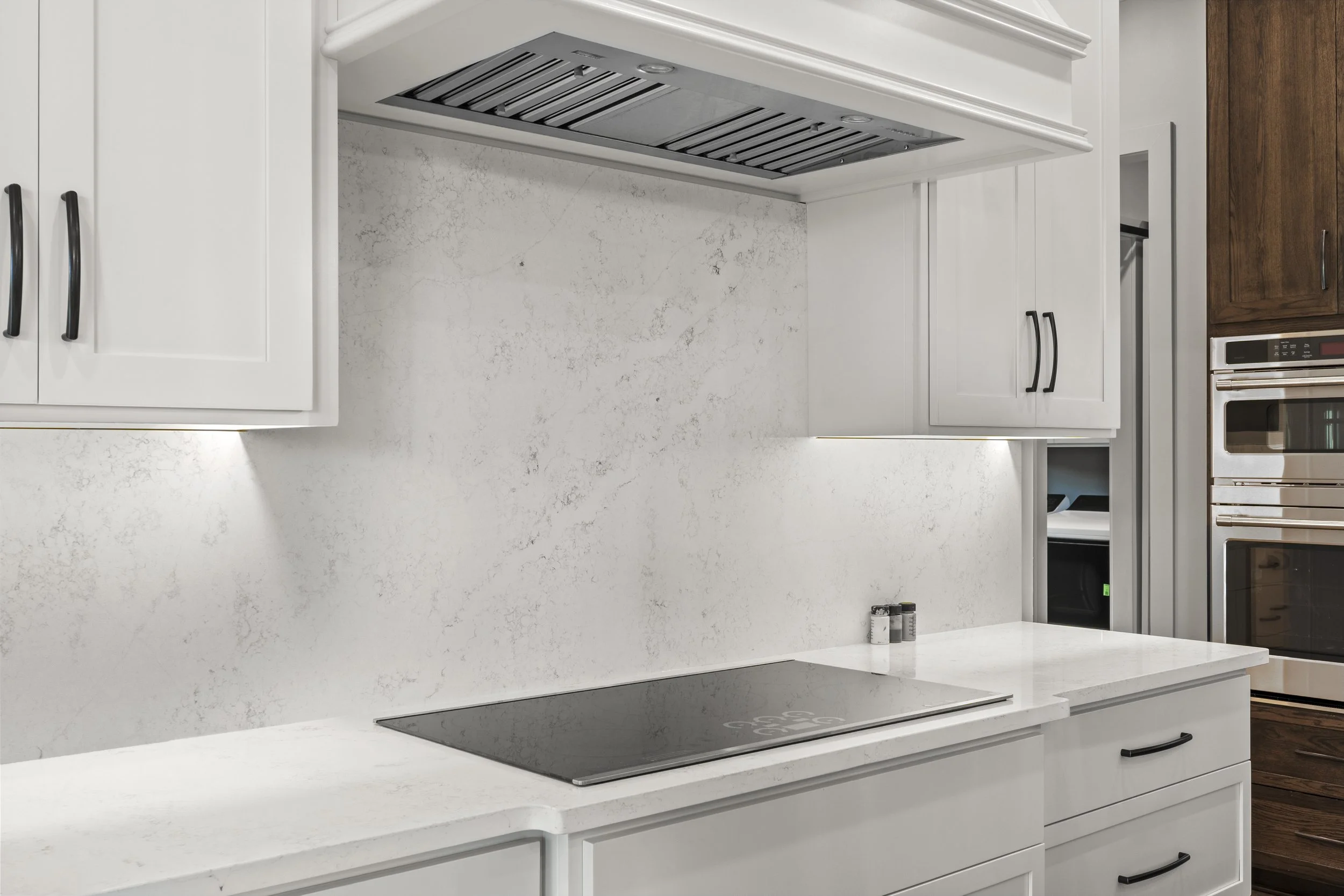 Modern kitchen with white cabinets, a gray marble backsplash, an induction cooktop, and a built-in oven. Wooden cabinets are visible in the background.
