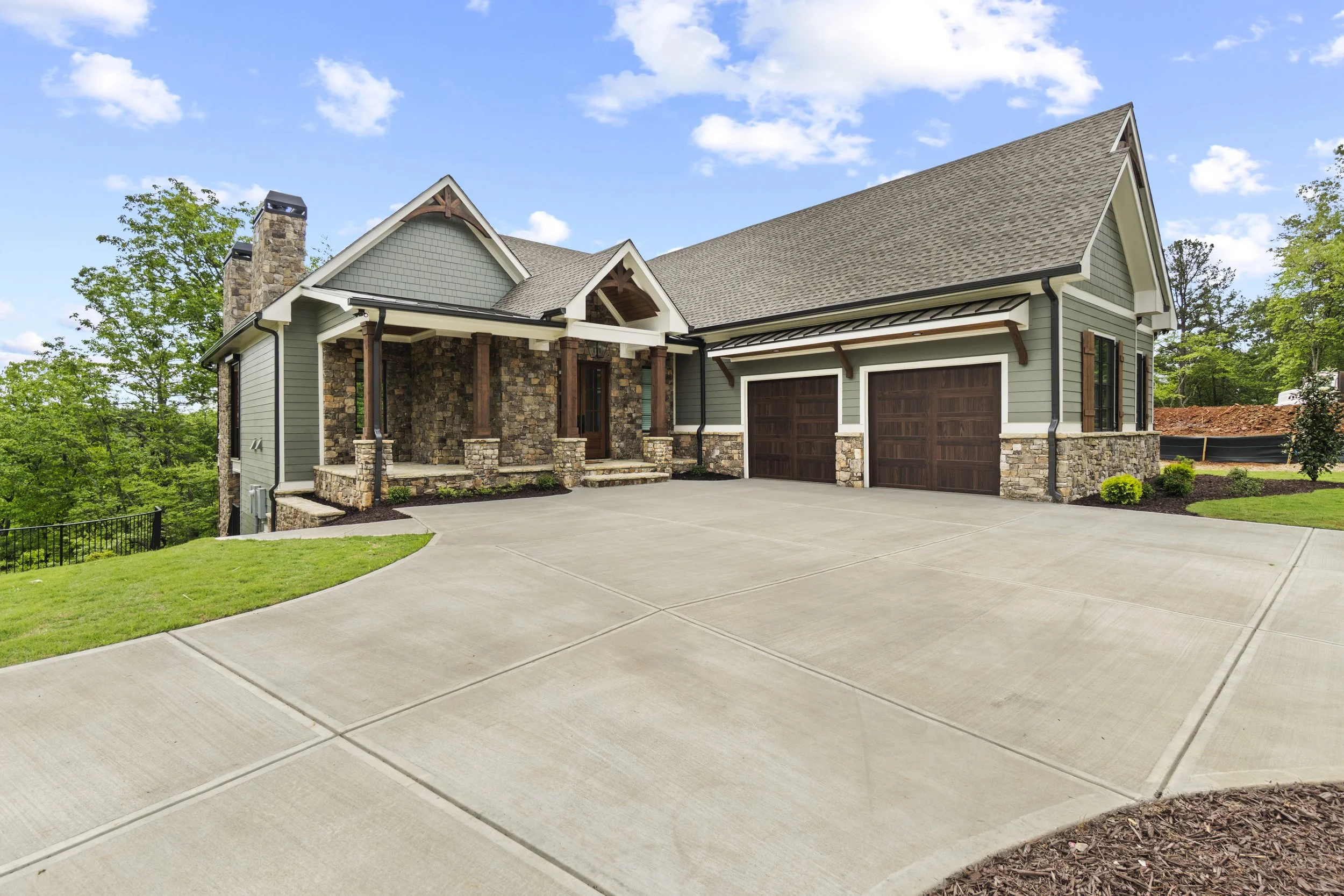 A modern house with a stone and siding exterior, a gable roof, and a two-car garage, surrounded by greenery and a spacious concrete driveway.