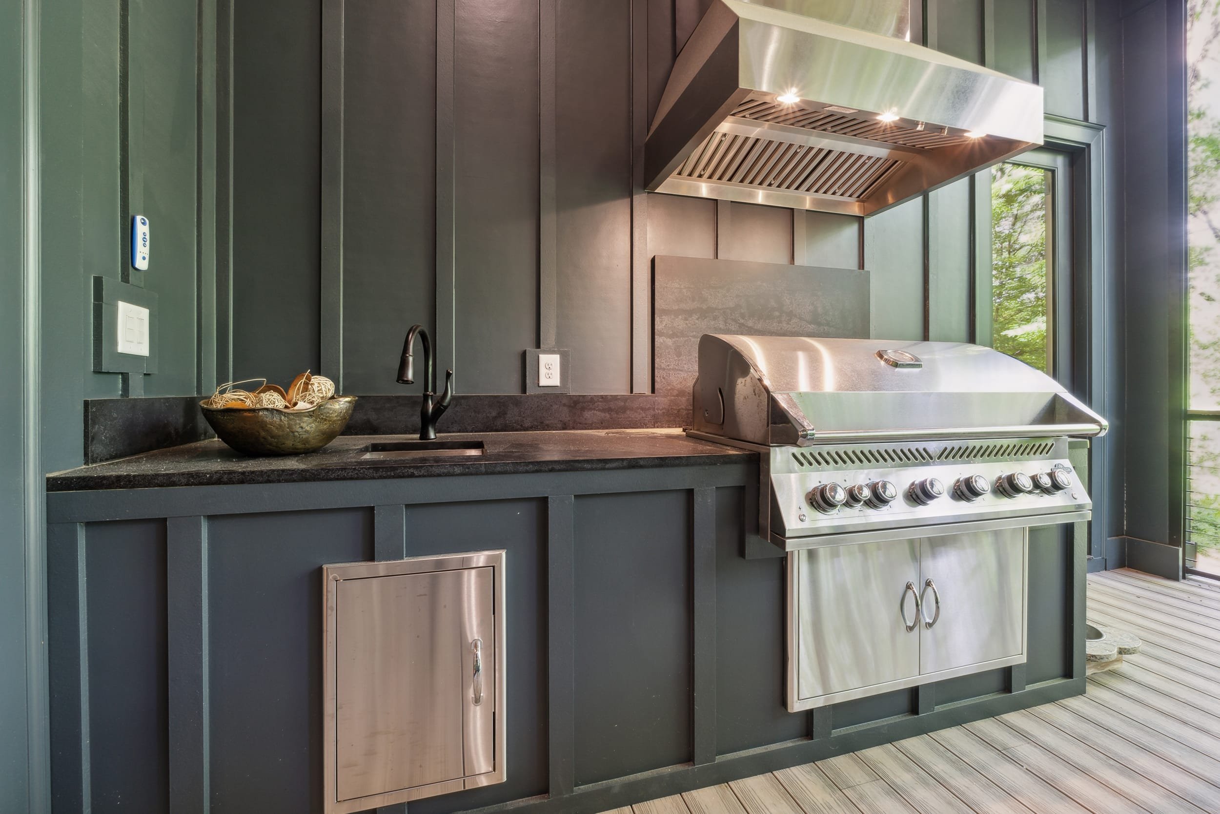 Modern outdoor kitchen with a stainless steel grill, black cabinetry, and a black countertop. A bowl with decorative balls sits next to a black faucet and sink. Large windows reveal greenery outside.