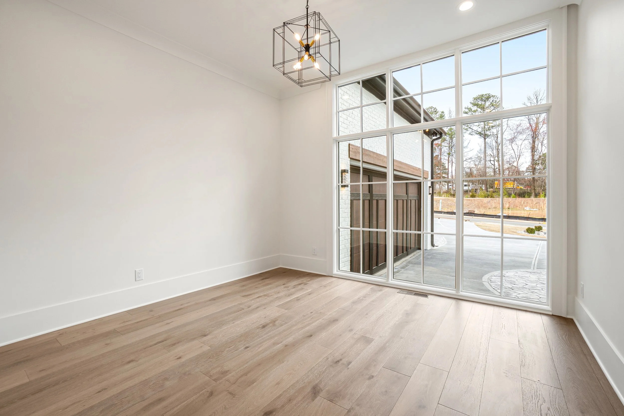 Empty room with large window, white walls, wood laminate flooring, modern black chandelier, outside view of trees and residential area.
