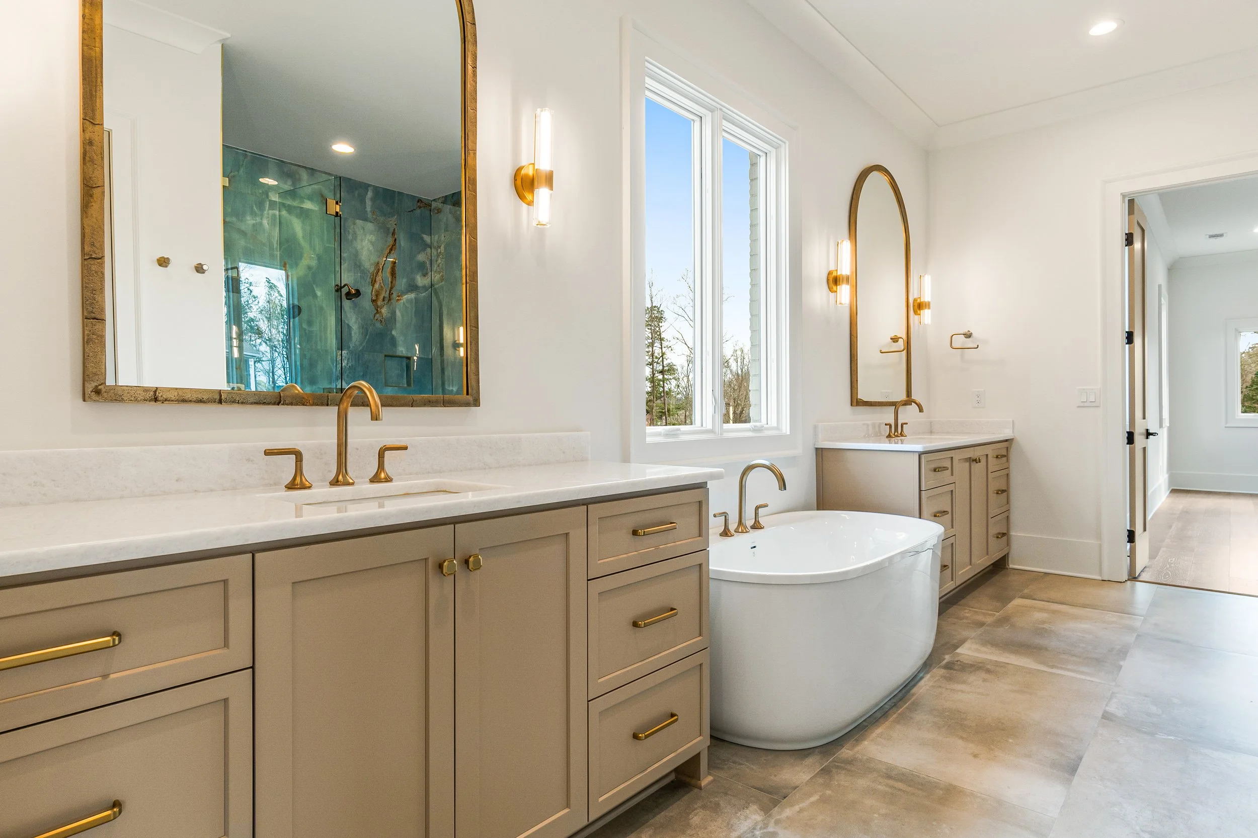 Modern bathroom with double vanity, large mirrors, two gold fixtures, natural light from windows, a freestanding bathtub, and a walk-in shower with glass doors and marble walls.