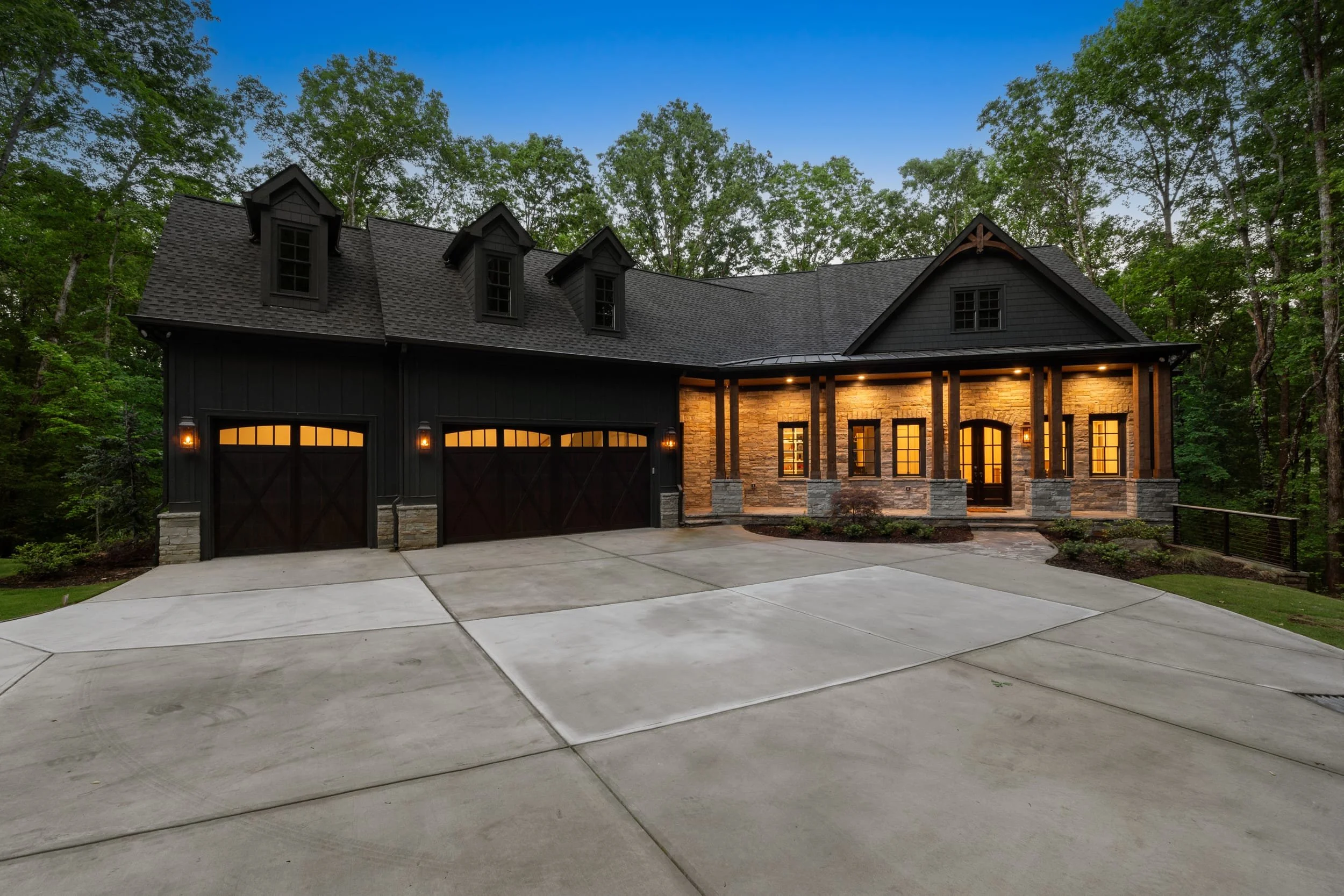 A modern house with a dark exterior, stone accents, four dormer windows on the roof, and warm exterior lighting, surrounded by trees and a wide concrete driveway.