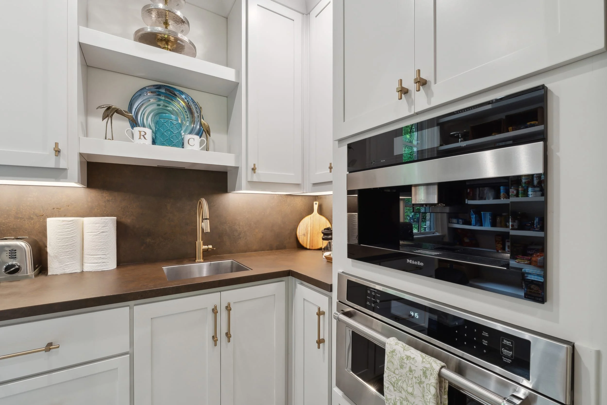 A modern kitchen with white cabinets, a black microwave oven, and a wood countertop with a sink. Decorative plates and cups are on the open shelves, and there's a toaster and paper towels on the counter. A cutting board leans against the wall, and a 