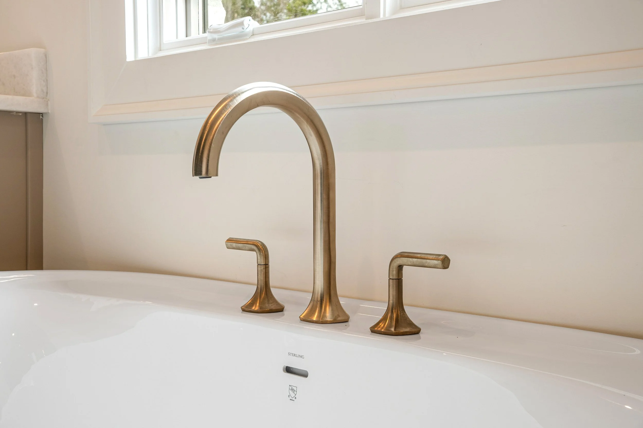 Close-up of a modern kitchen sink with a brushed gold faucet, set against a white countertop and wall, with a window in the background.