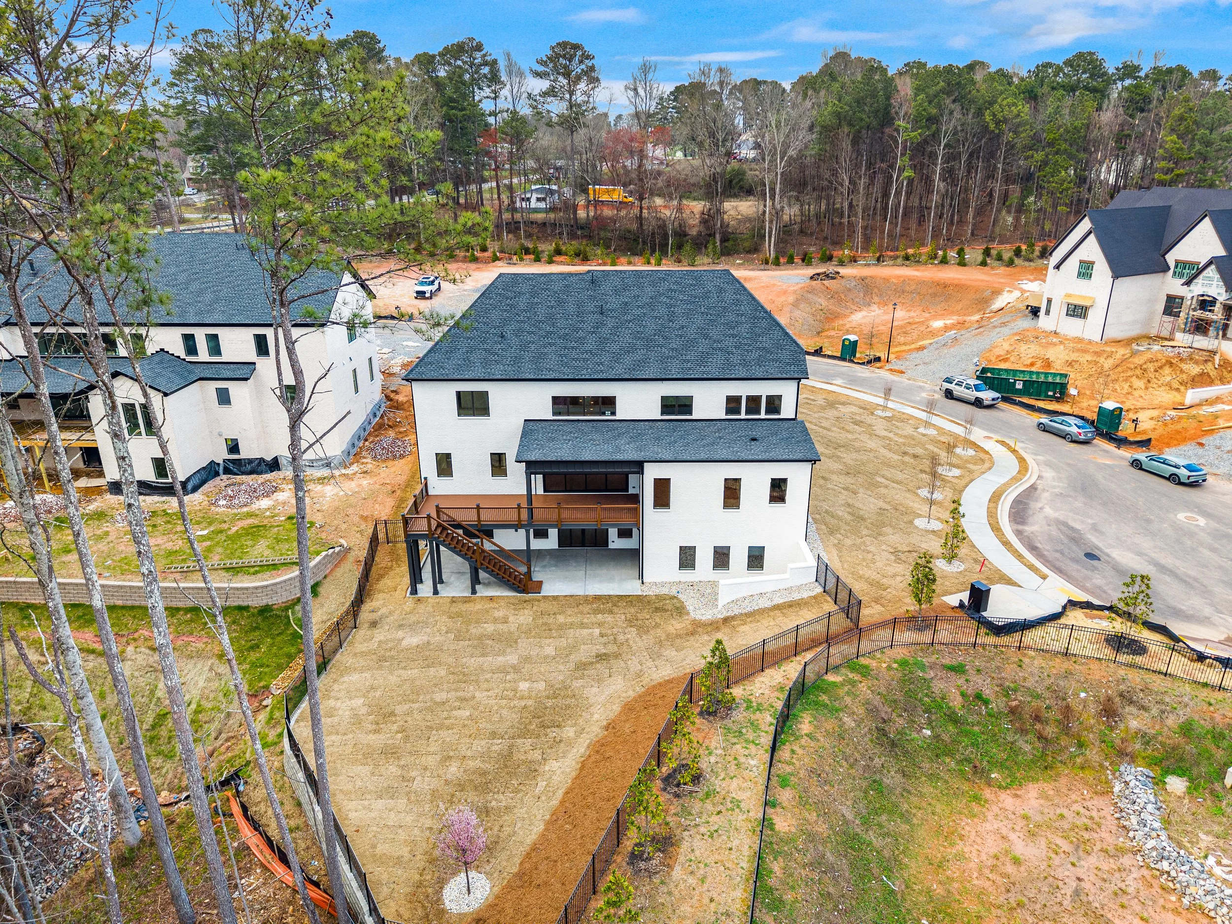 Aerial view of a residential construction site with partially built houses, a paved terrace, small trees, and a curved driveway, surrounded by green and brown trees under a partly cloudy sky.