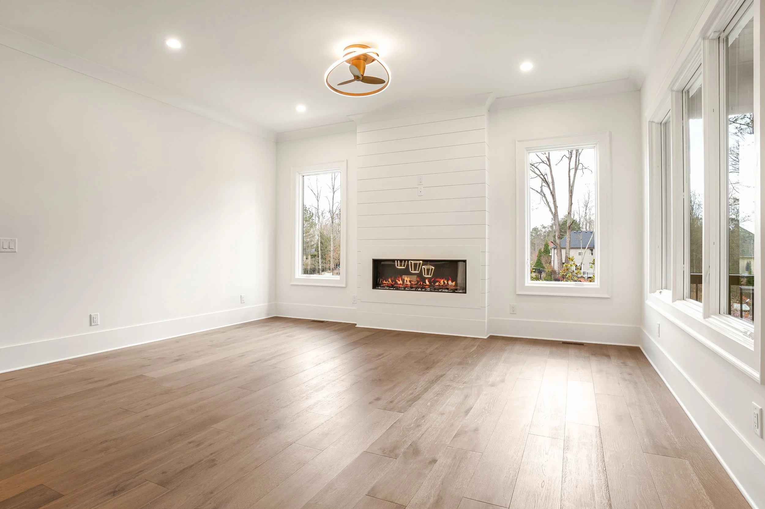 Empty living room with white walls, four large windows, a fireplace, a ceiling fan, and hardwood floors.