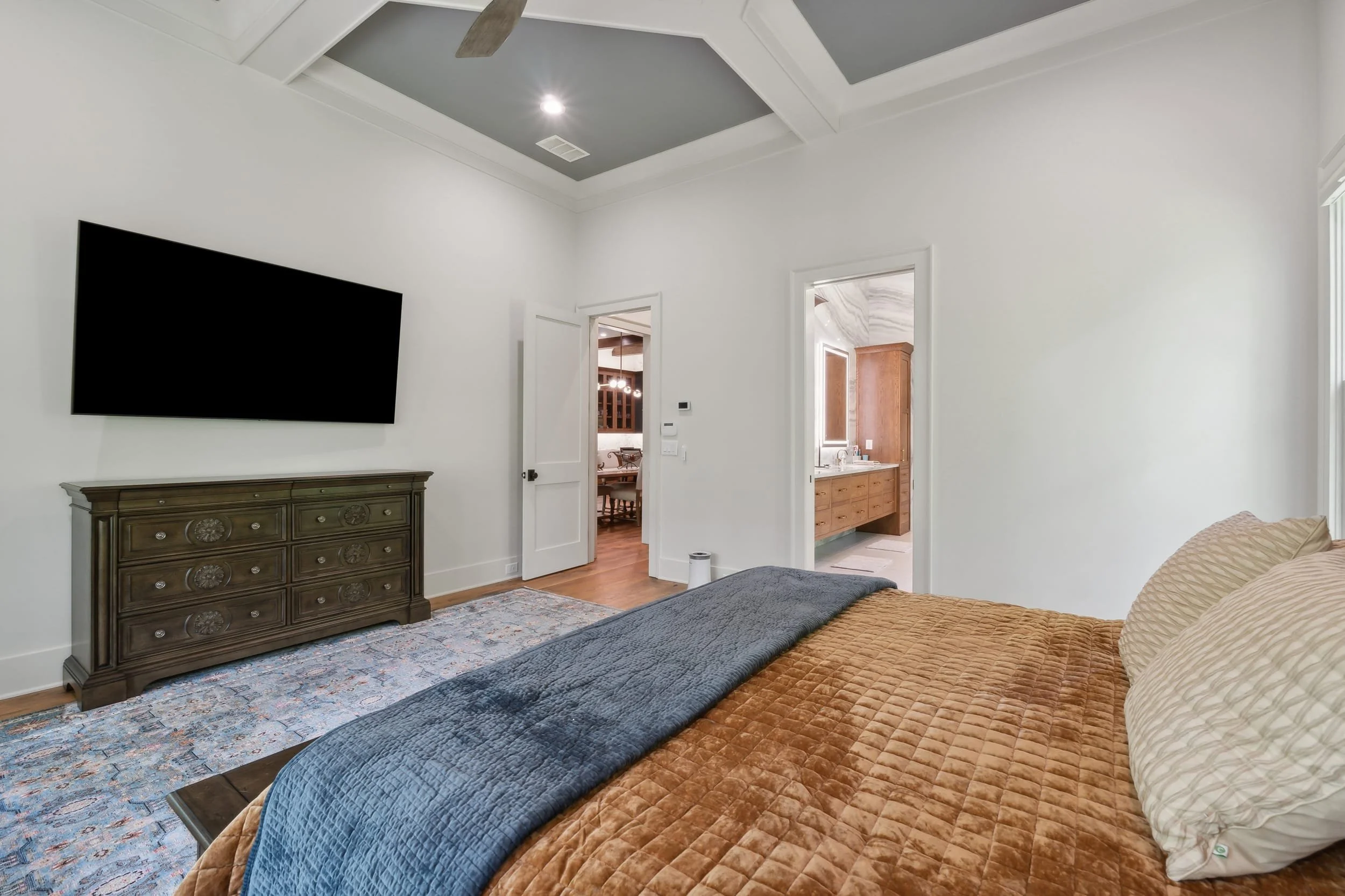 A bedroom with a bed covered in a brown quilt, a blue quilted throw blanket, and pillows. A vintage wooden dresser is against a white wall, below a large flat-screen TV. The room has white walls, a patterned area rug, and a ceiling with coffered desi