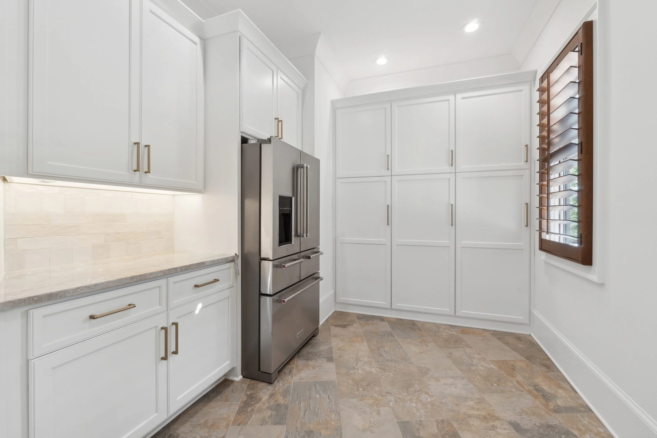 A bright kitchen with white cabinets, a stainless steel refrigerator, and a window with wooden blinds.