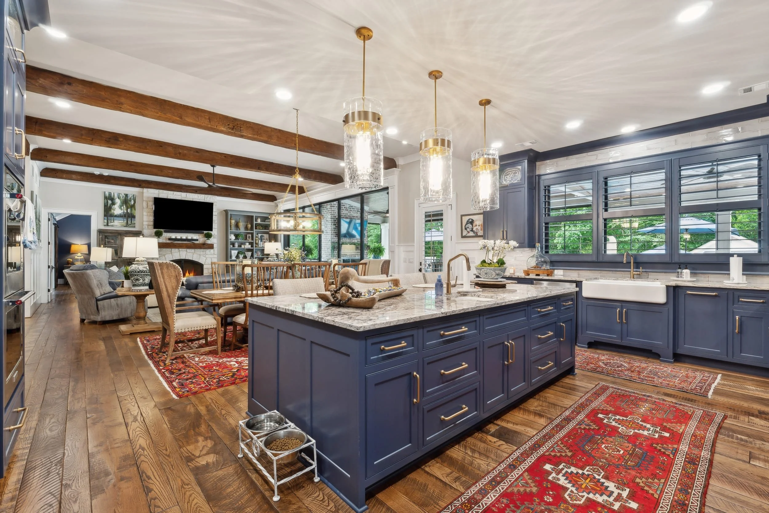 Open concept kitchen and living room with blue cabinets, granite countertops, wood flooring, and exposed wooden beams on the ceiling. Pendant lights hang over the kitchen island, which has a white farmhouse sink. The living room features a fireplace, a large flat-screen TV, and various furniture. Large windows with shutters let in natural light and show greenery outside.