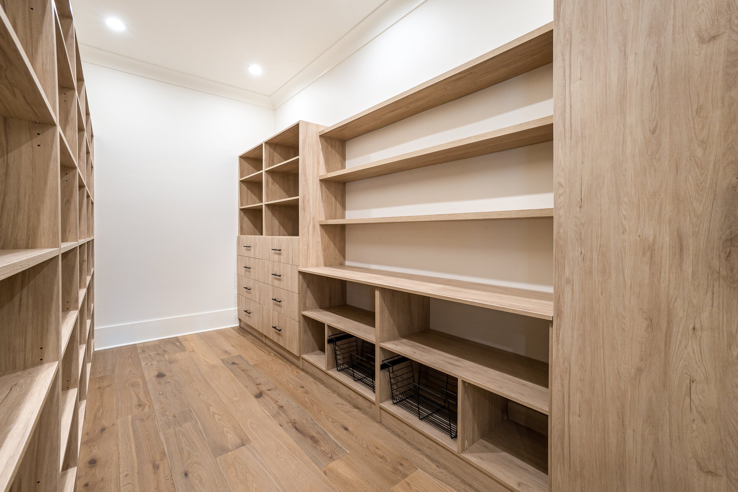 Empty wooden closet with open shelves and drawers, hardwood floor, white walls, and recessed ceiling lighting.