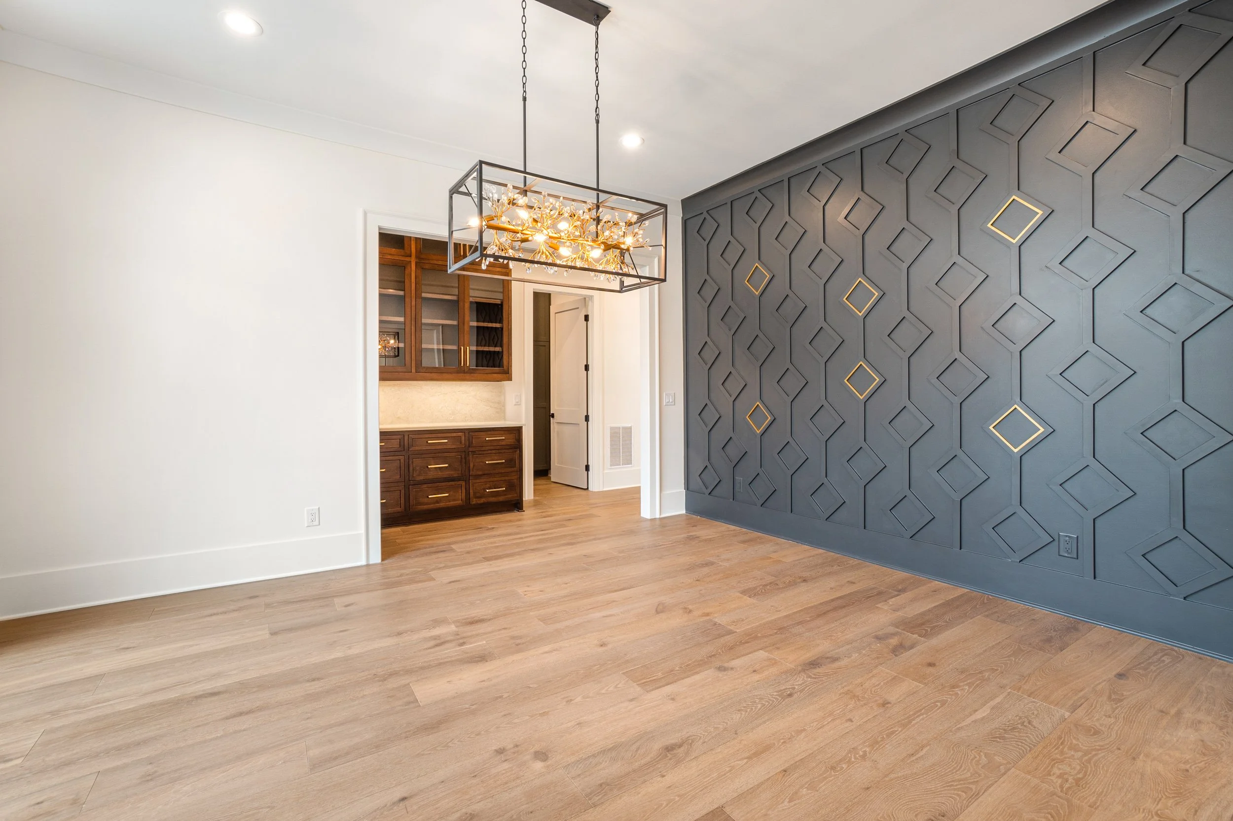 Interior of a modern home with a hardwood floor, a gray accent wall with geometric patterns, a chandelier, and a small built-in wooden cabinet with glass doors.