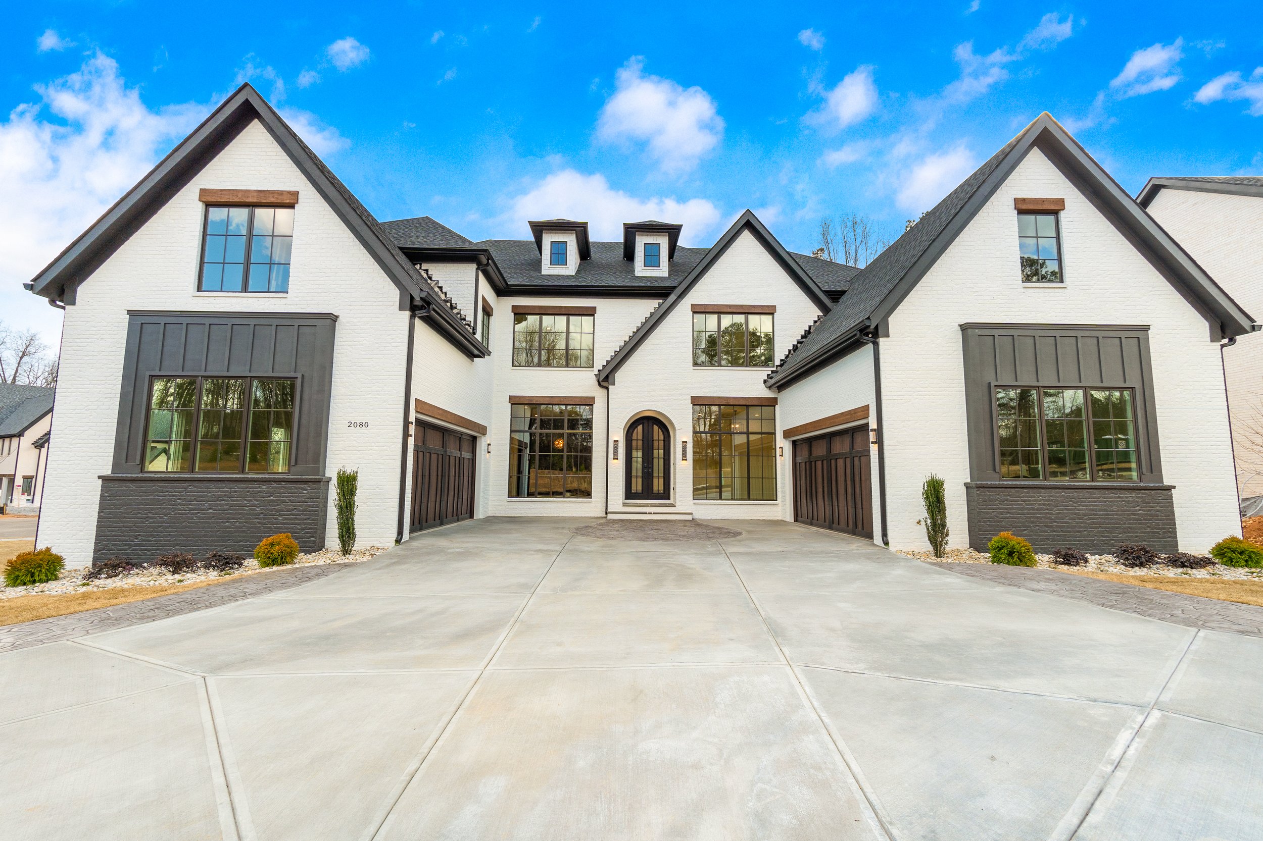 A large, modern white house with a black roof, large windows, and a double garage, situated under a bright blue sky with some clouds.
