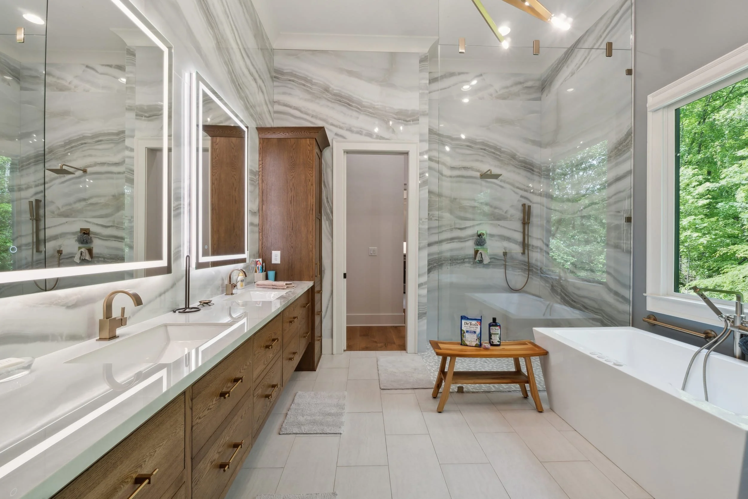 Modern bathroom with marble walls, a double vanity with lighting, wooden cabinets, a large window with greenery outside, a soaking tub, and natural light.