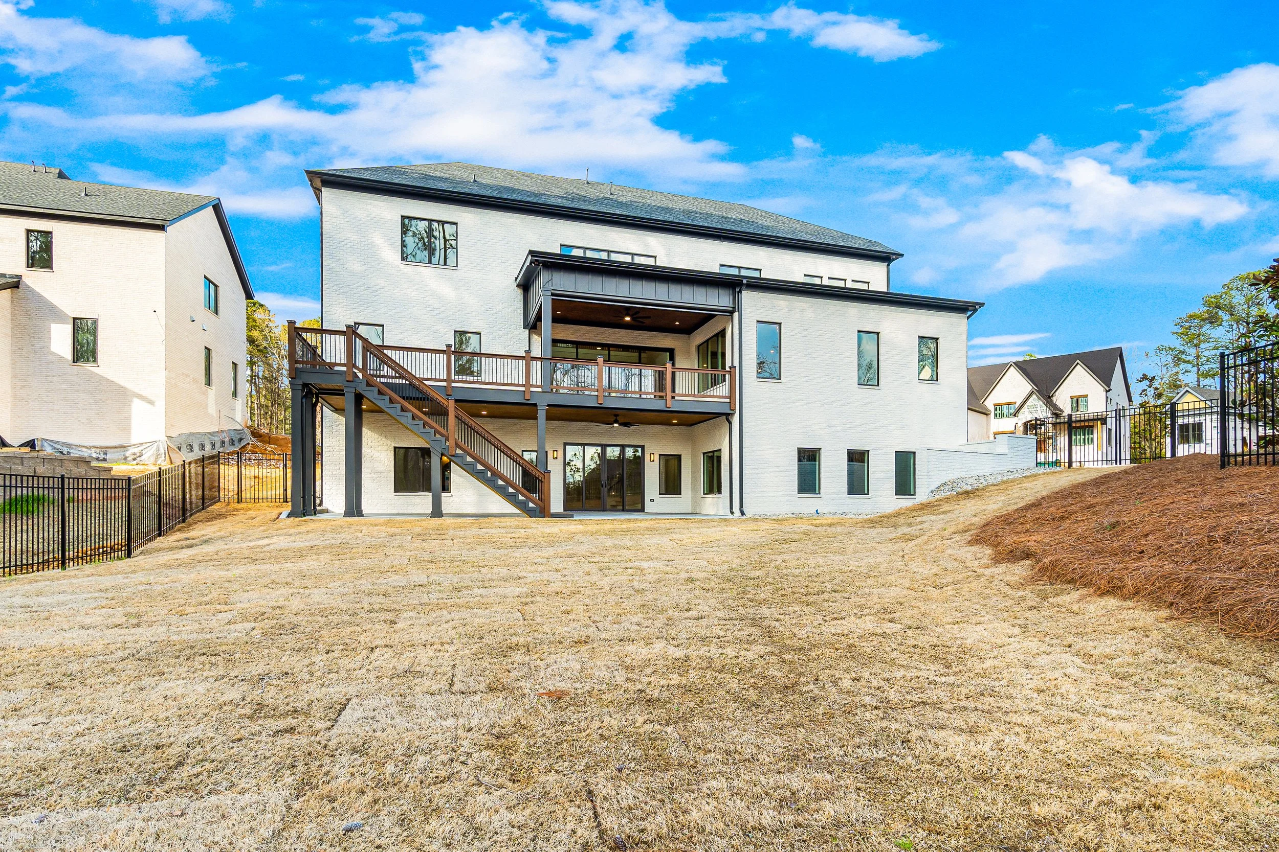 A modern two-story house with white brick exterior, large windows, and a wooden deck with stairs, surrounded by a yard with dry grass and a black fence, under a partly cloudy blue sky.