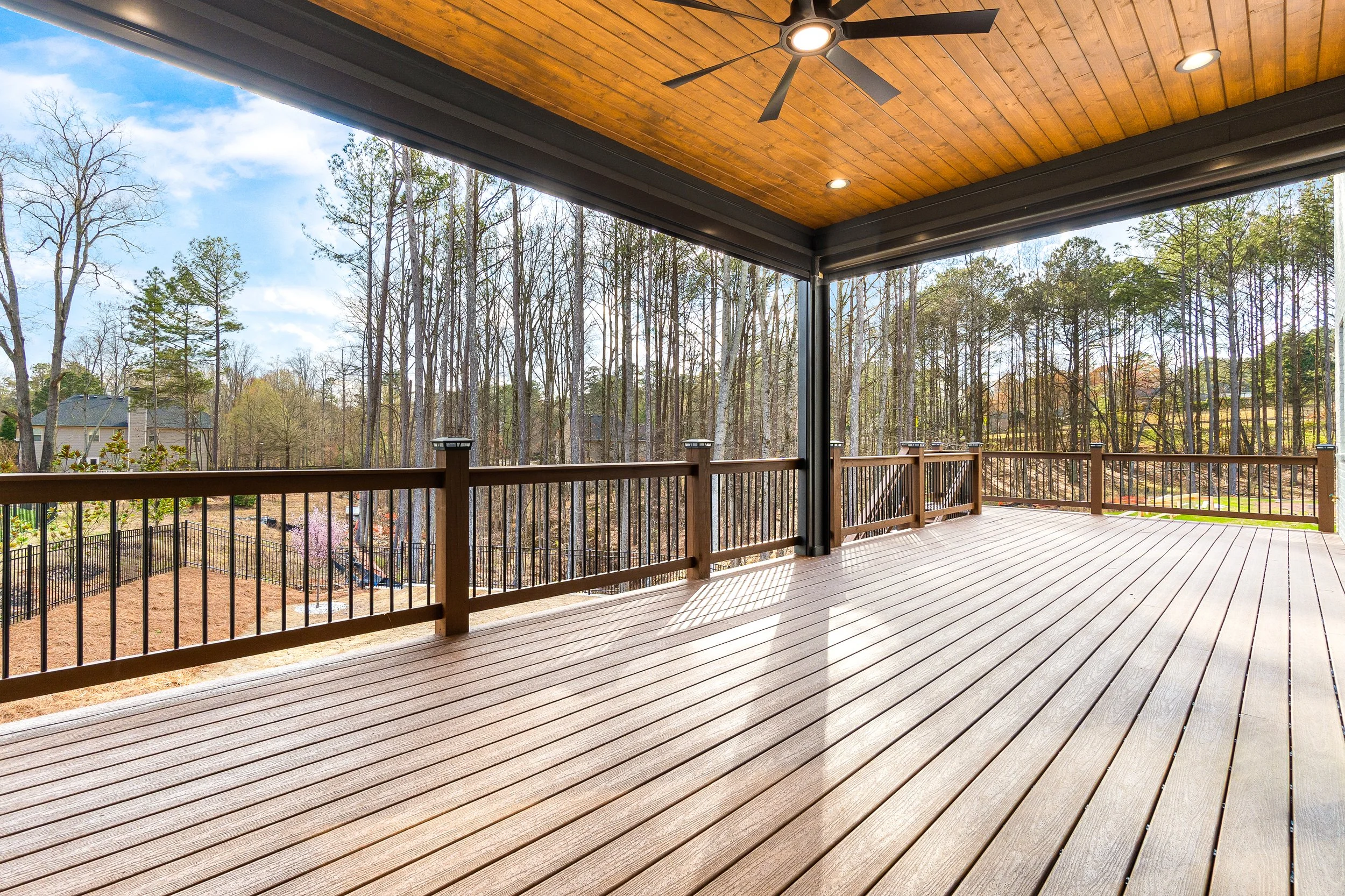 A spacious wooden deck with a black railing, overlooking a wooded area with tall trees and a few houses in the background. The ceiling of the porch is wooden with recessed lighting and a ceiling fan.