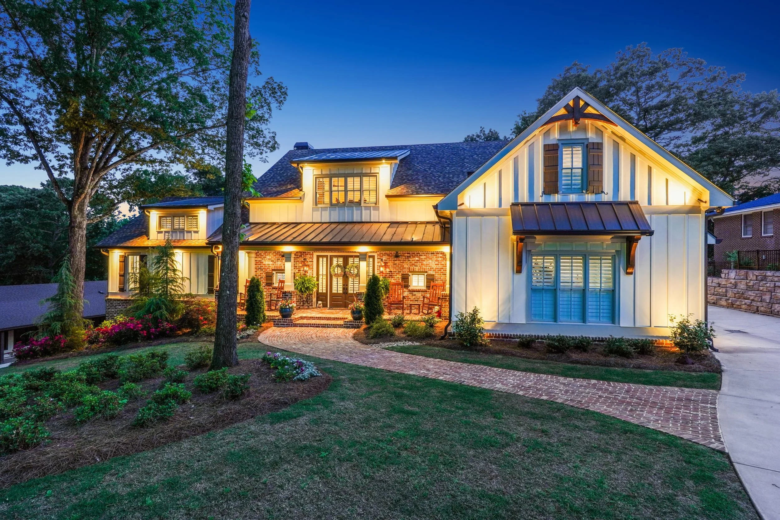 A two-story house illuminated at dusk surrounded by trees, with a brick pathway leading to the front entrance and landscaped yard with flowers.