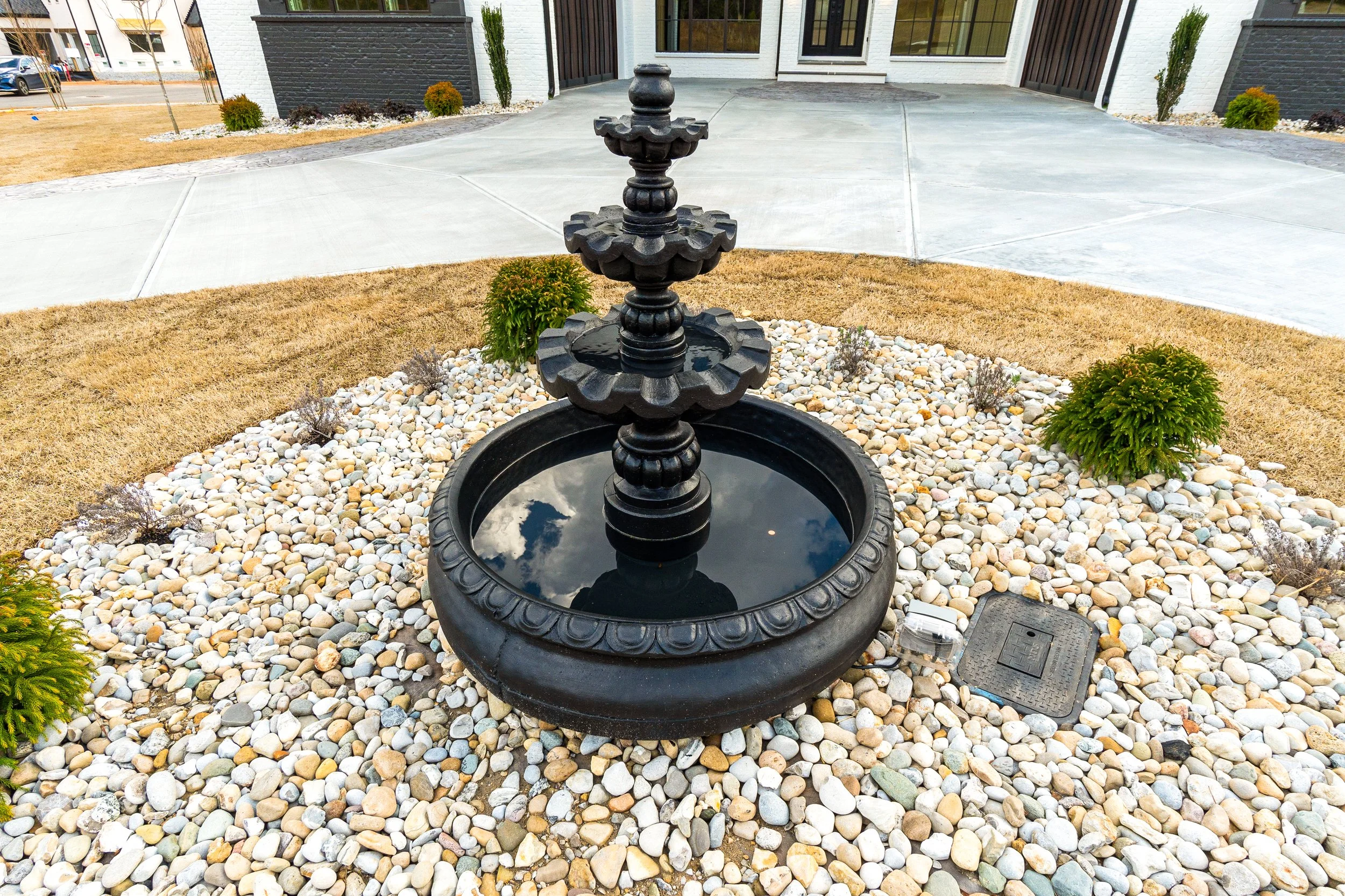 Black decorative tiered water fountain in a landscaped yard, surrounded by small rocks, with a house and parked cars in the background.