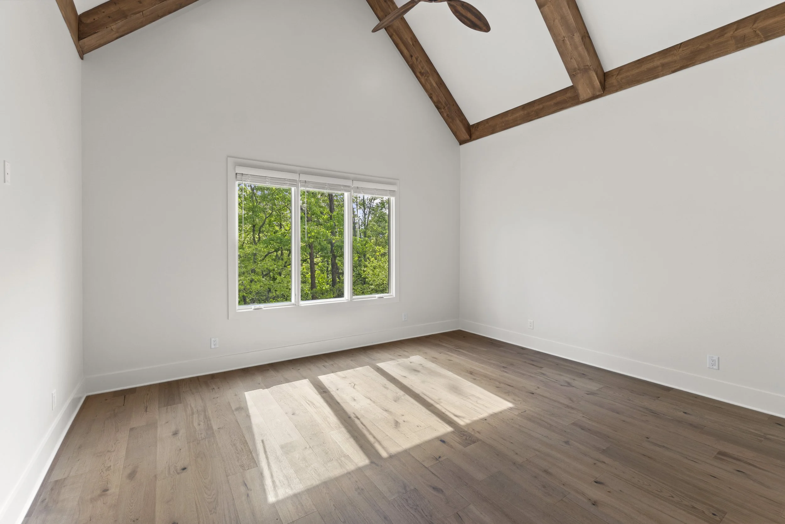 Empty room with white walls, a window with blinds, wooden flooring, and exposed wooden beams on the ceiling, with greenery visible outside the window.