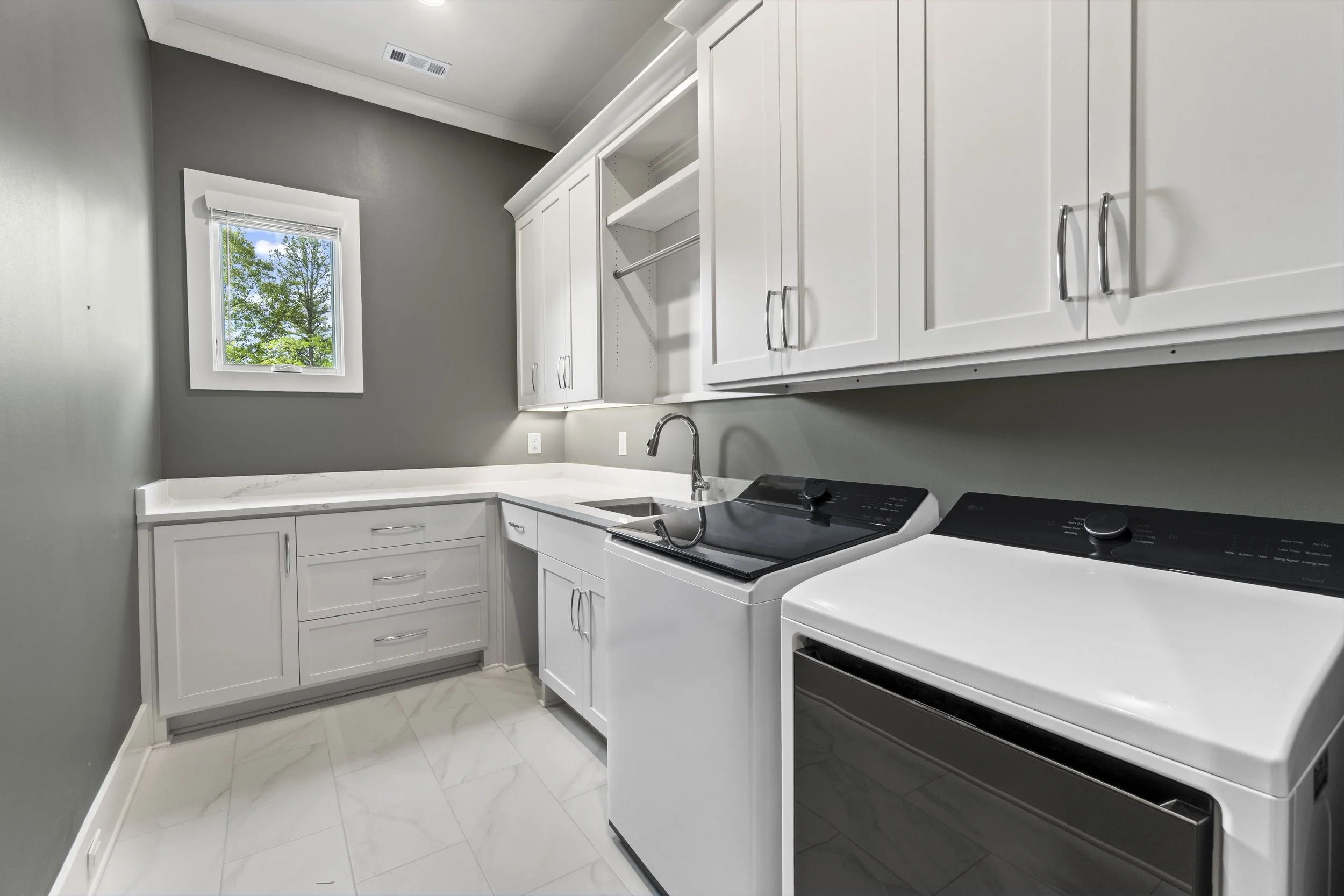 A modern laundry room with white cabinets, a white countertop, a stainless steel faucet, a washing machine, and a window showing green trees outside, painted in gray.