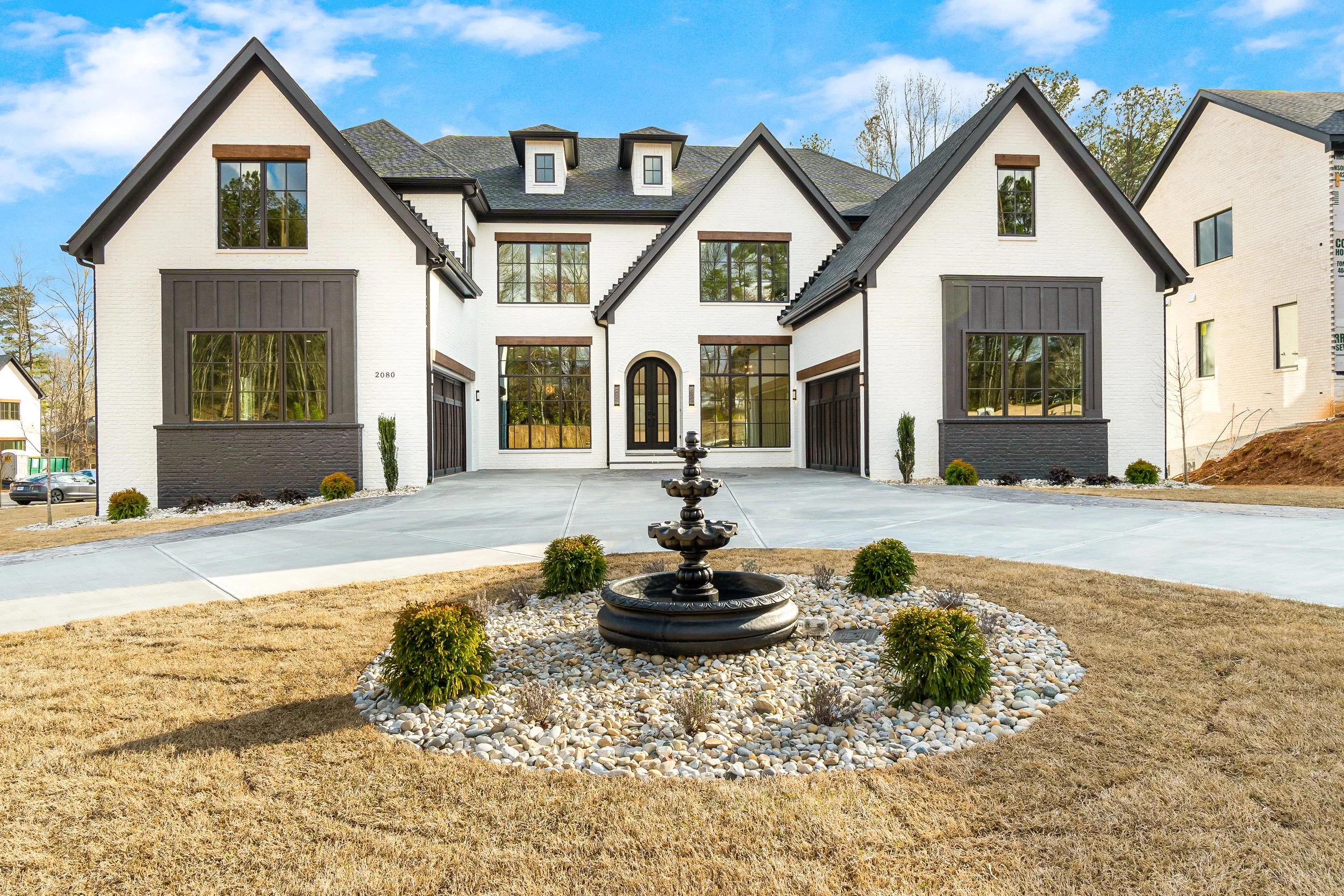 Front view of a modern white house with black accents, large windows, a driveway, a fountain, and small landscaped bushes, under a blue sky.