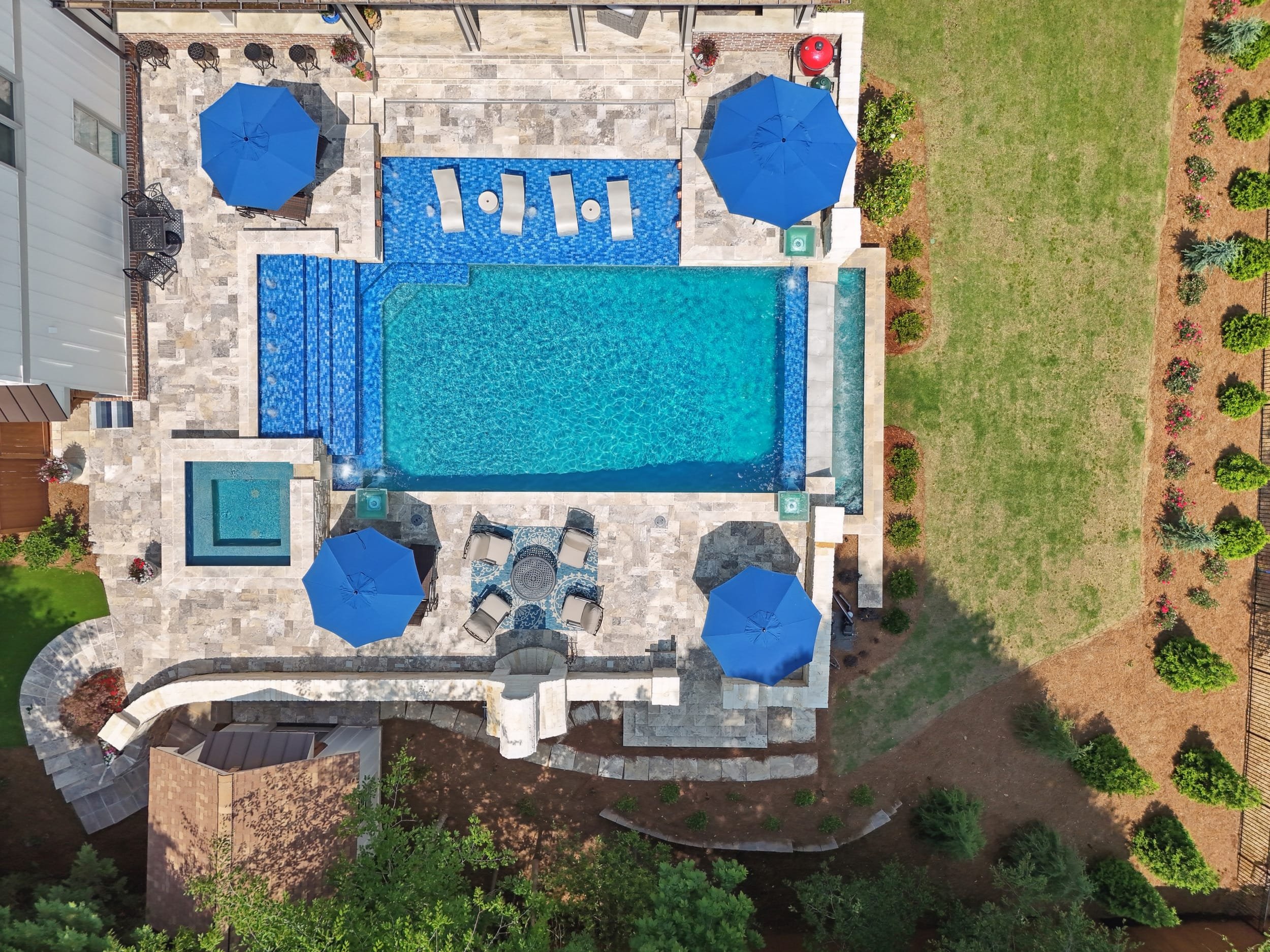 Top-down view of a backyard with a swimming pool, two blue umbrellas, patio furniture, and landscaped grass area with bushes and flowers.