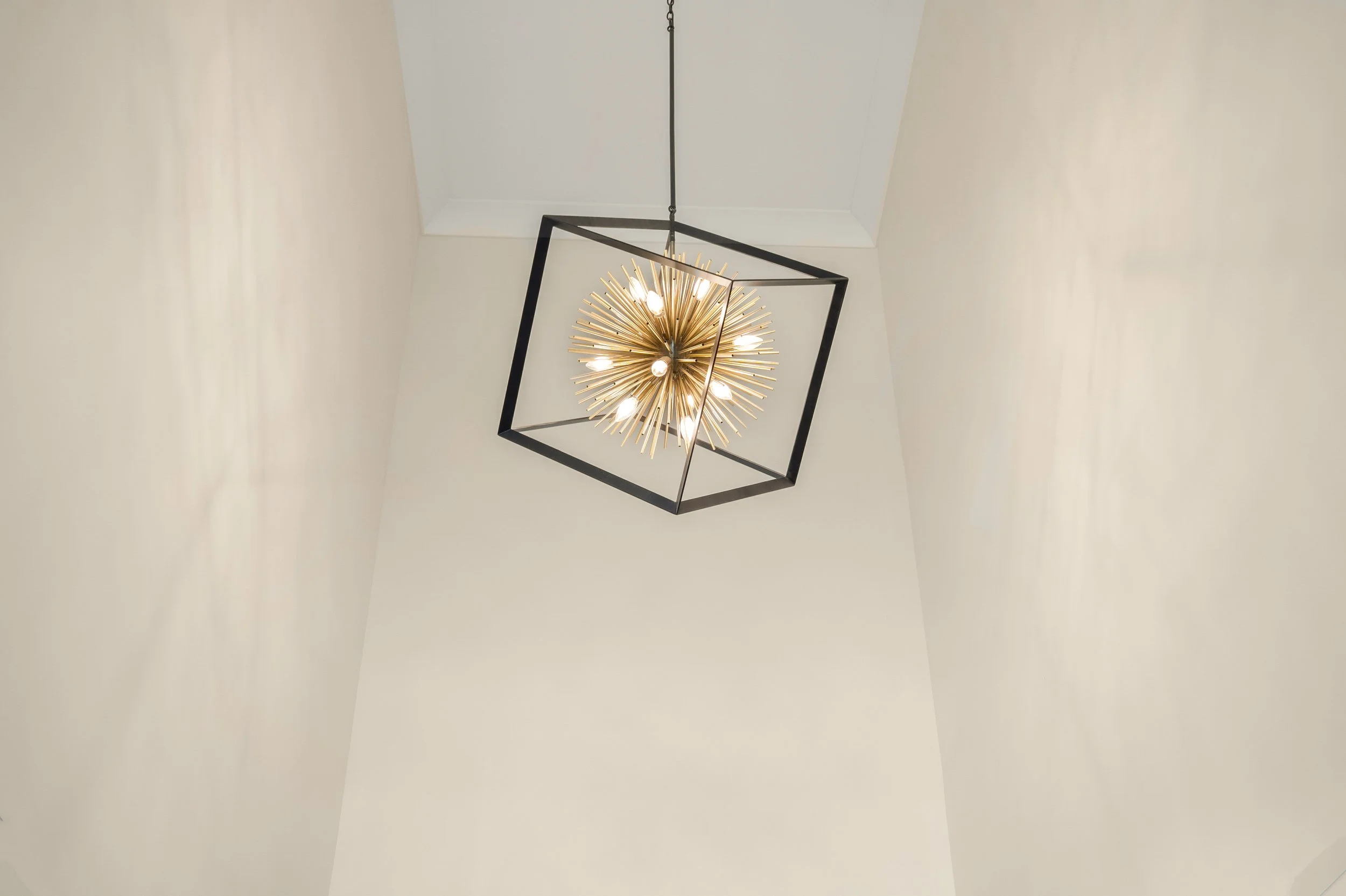 Modern chandelier with multiple bulbs and gold spokes inside a black square frame, hanging from a high ceiling.