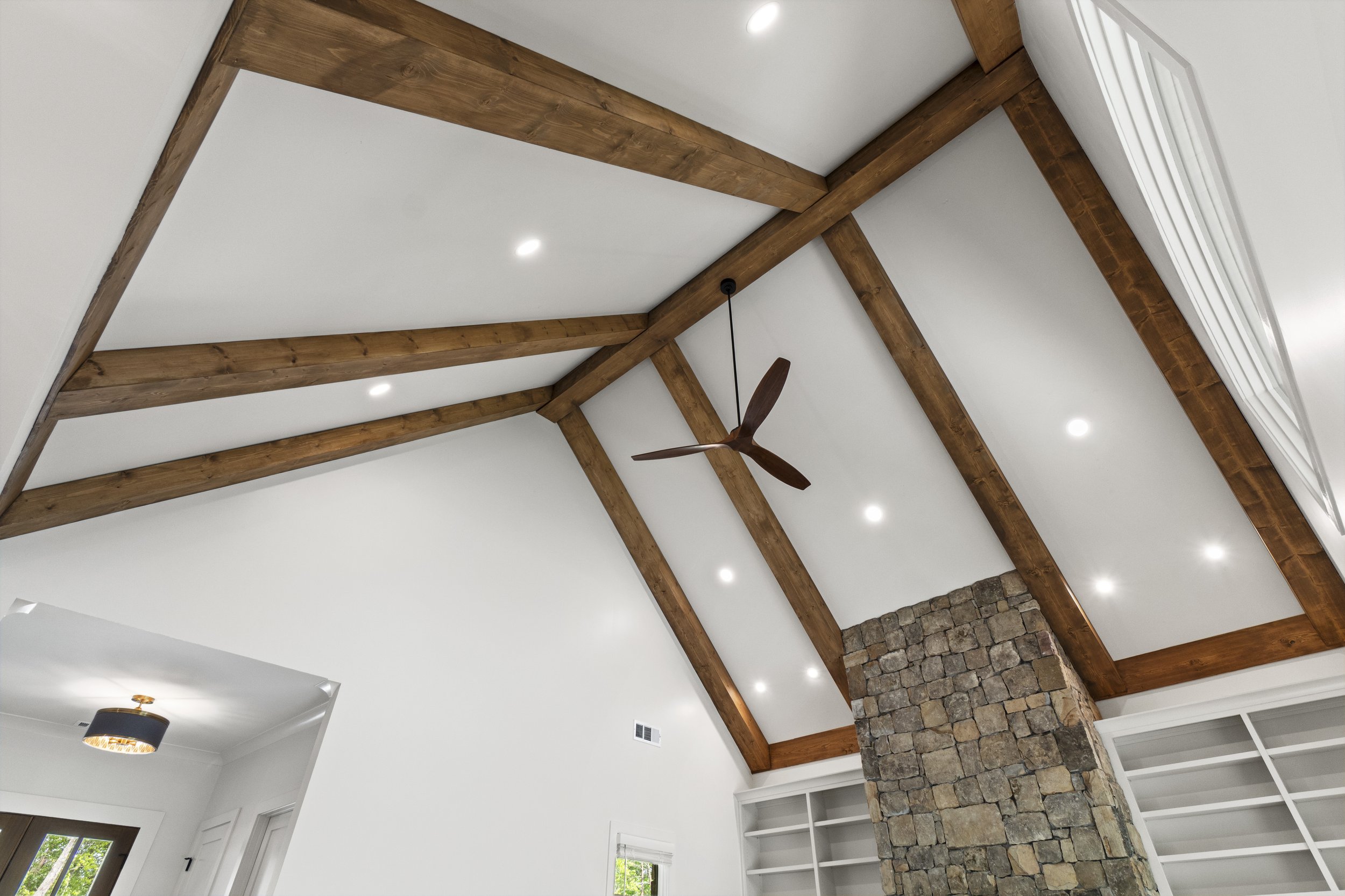 View of a room with white ceiling and walls, wooden beams in a vaulted ceiling, a ceiling fan, and recessed lighting. Part of a stone fireplace and built-in shelves are visible.