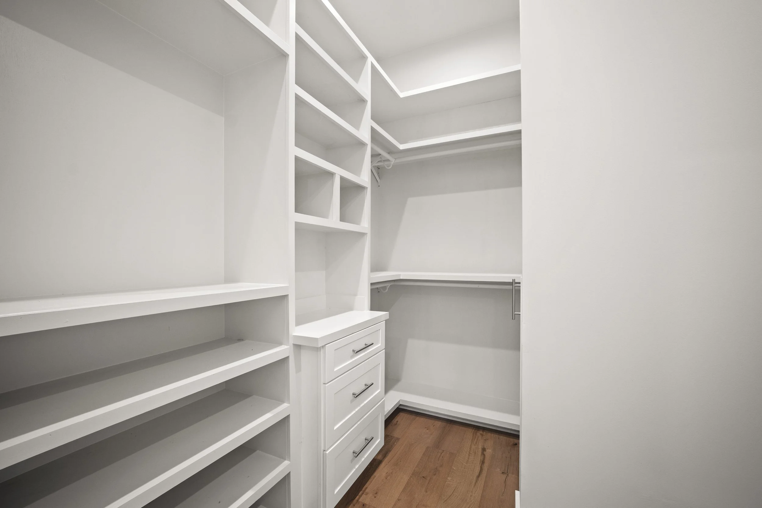 Empty white walk-in closet with built-in shelves, drawers, and hanging space, and hardwood floors.