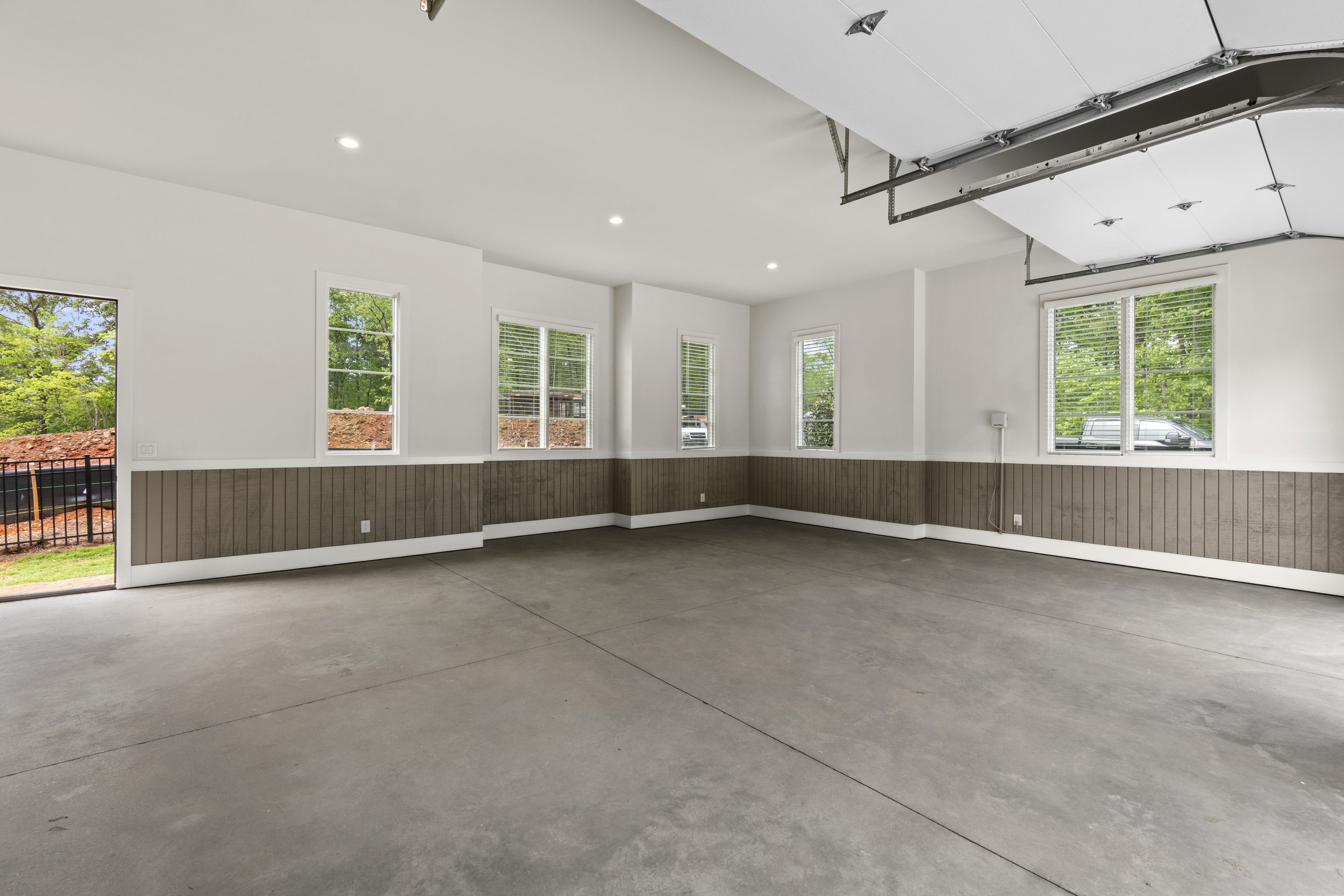Unfinished garage with white walls, brown wainscoting, concrete floor, and a partly open garage door showing trees and a parked vehicle outside.
