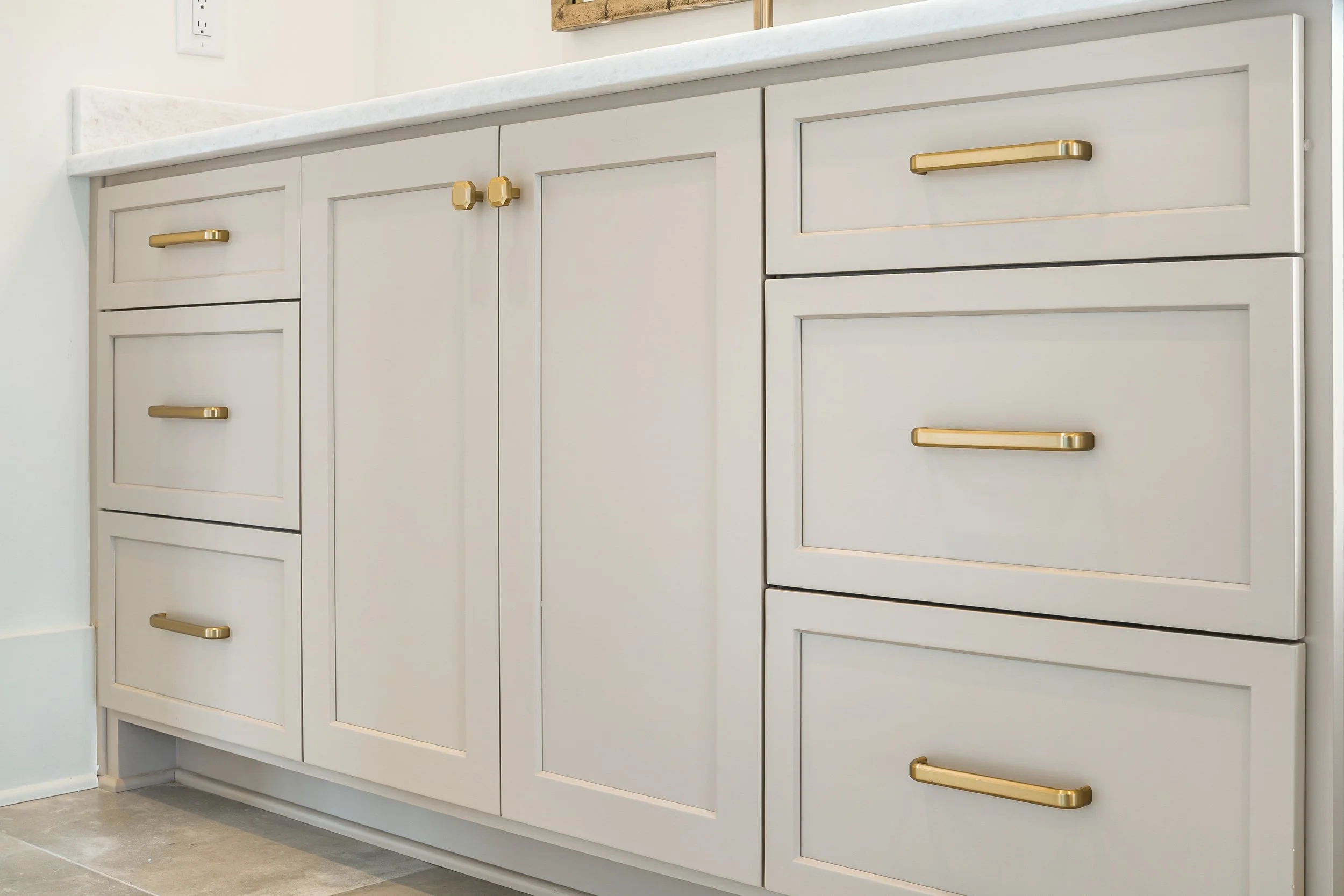 White kitchen cabinet with gold handles, marble countertop, next to a white wall with an electrical outlet.